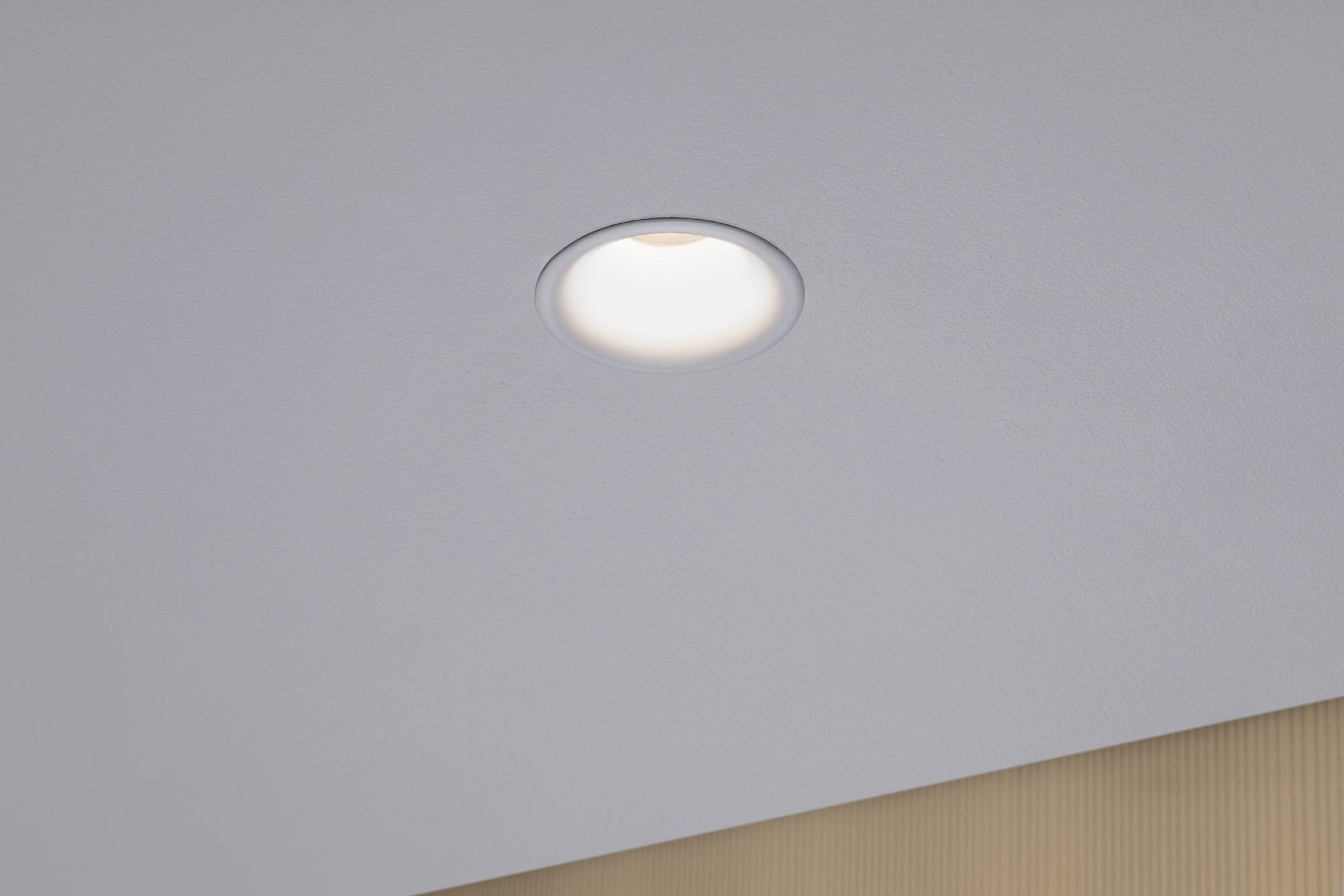 Ein eingelassener Deckenstrahler ist in der Mitte einer weißen Decke zentriert. Licht geht von der Mitte der Leuchte aus.