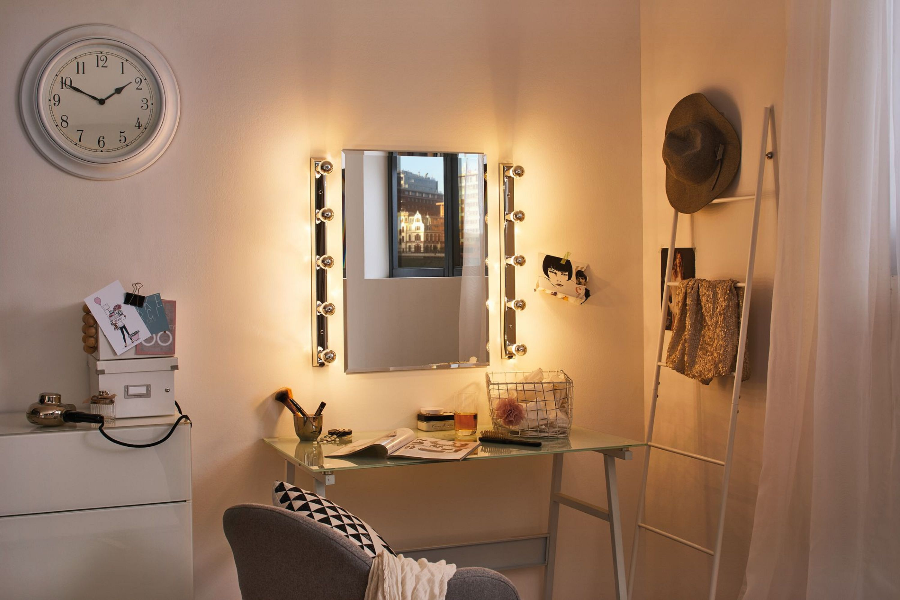 Toaletka z lustrem, światłami i biurkiem. Zegar wisi nad nią. Kapelusz i koc na drabinie.