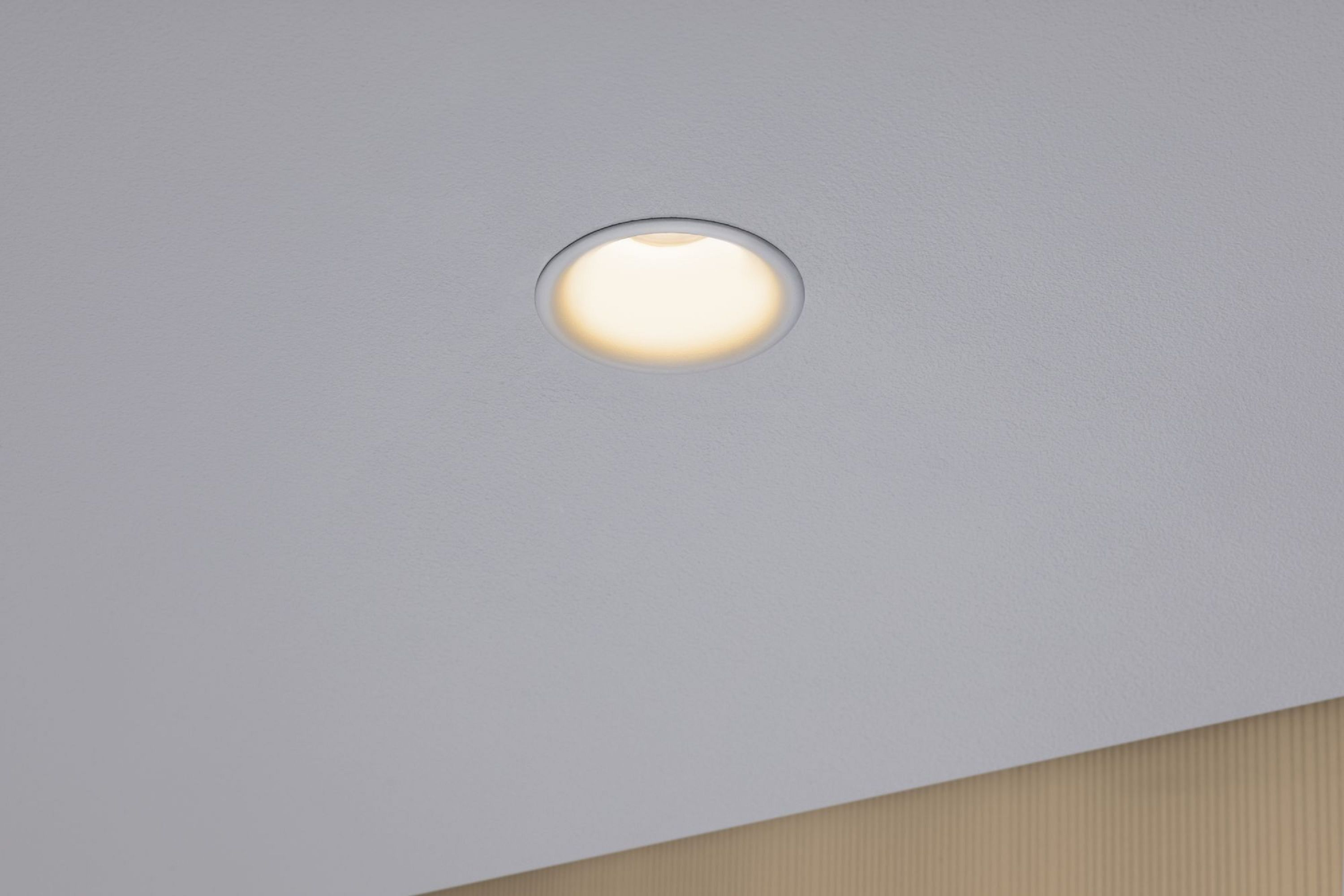 Un plafond avec un luminaire encastré. La lumière est circulaire et vive. Le plafond est blanc avec un coin beige clair.
