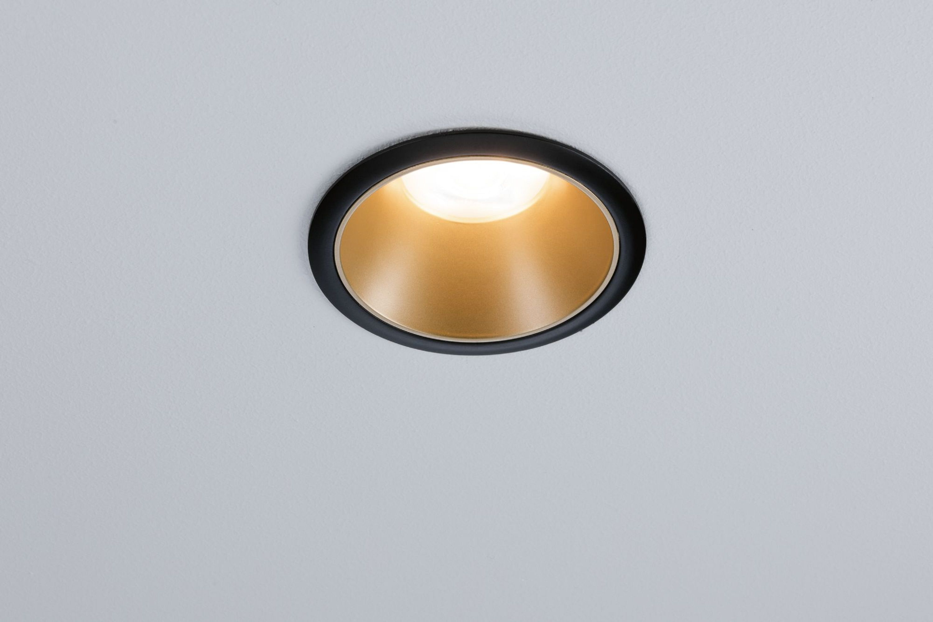 Un faretto da incasso. Ha un anello nero, interno dorato ed emette una luce intensa su soffitto bianco.