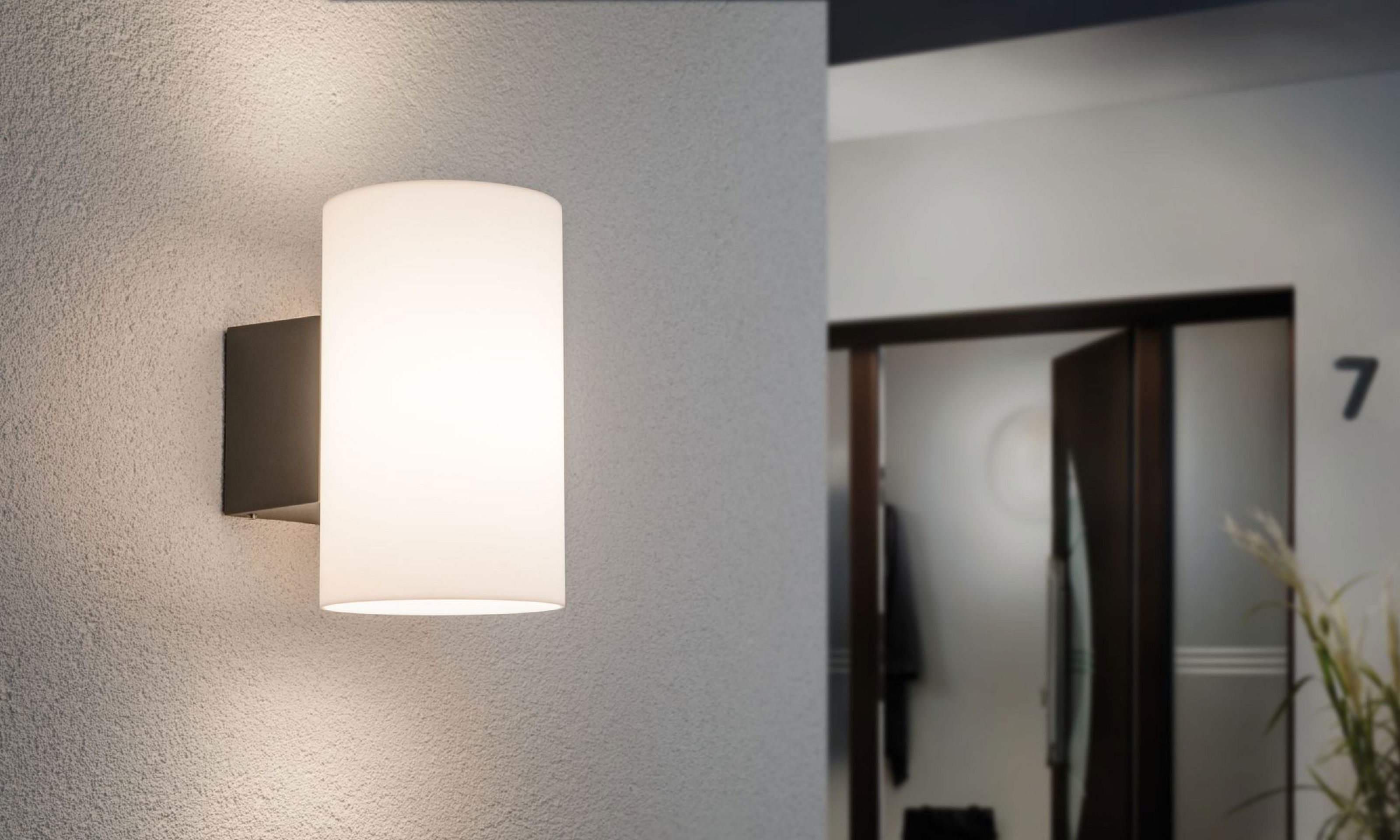 Cylindryczna lampa ścienna z białym kloszem jest zamontowana na teksturowanej szarej ścianie. Tło pokazuje otwarte drzwi.