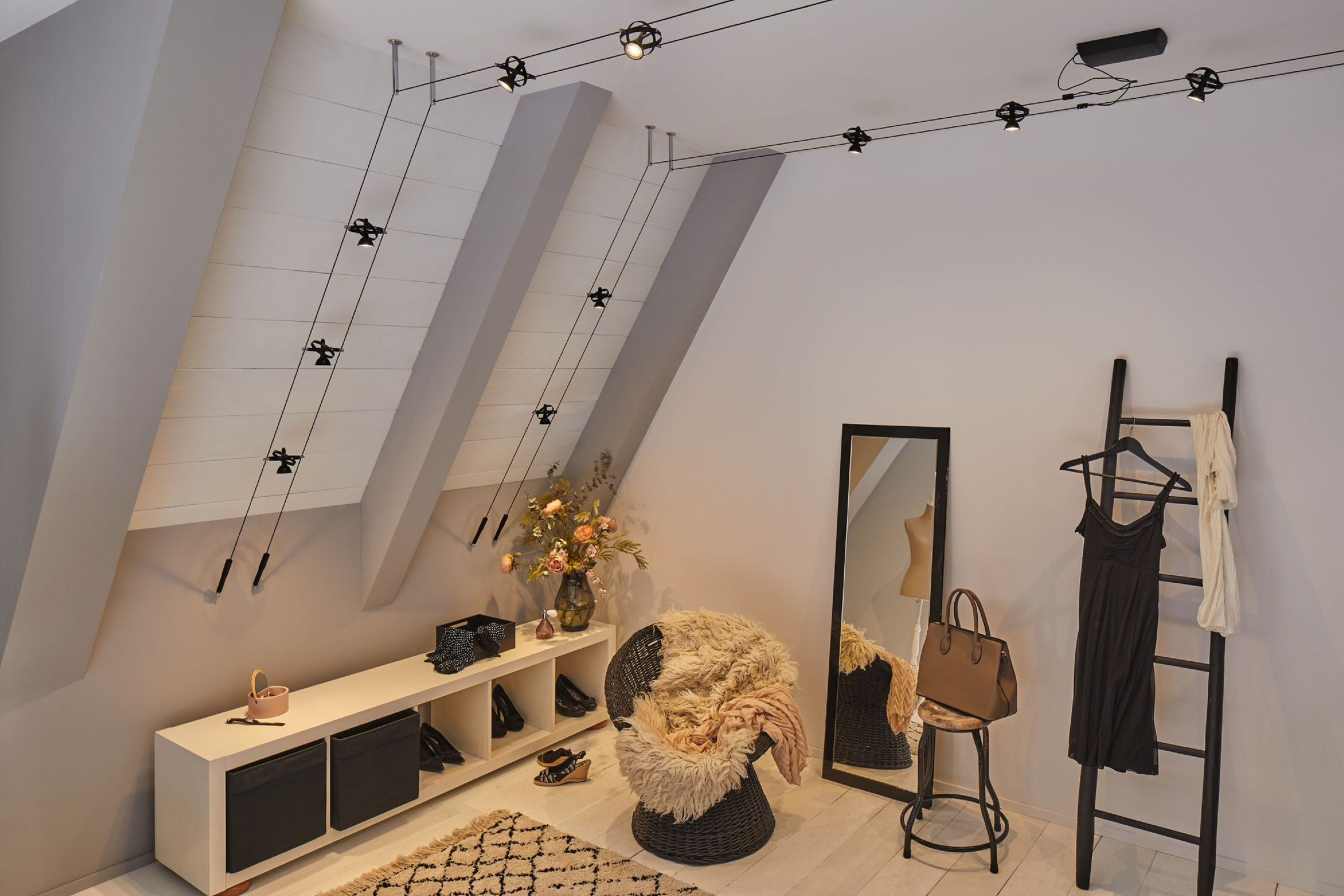 Una stanza con soffitto spiovente, illuminazione a binario, specchio, vestiti, scala, sedia con pelliccia e scarpiera.