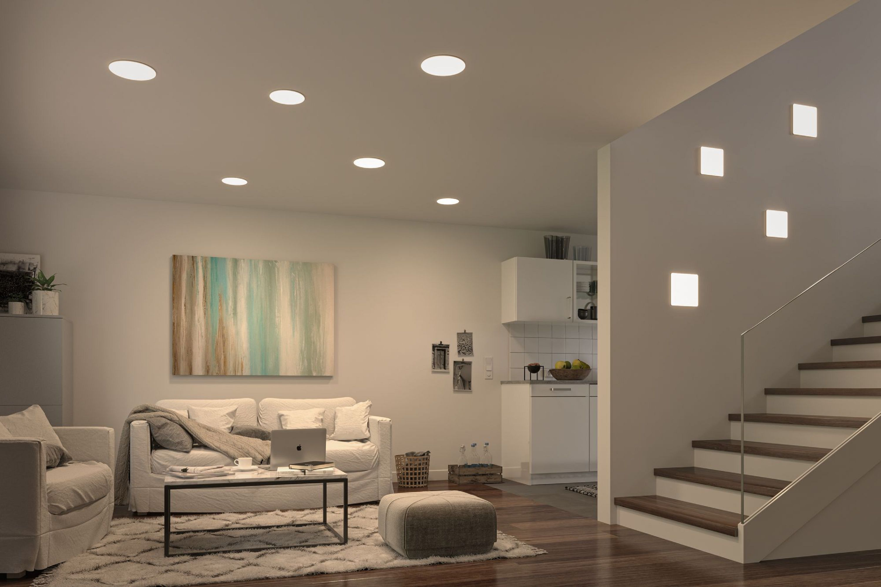 Soggiorno con divano, tavolino e arte astratta. Sono visibili luci a soffitto e scale. Cucina sullo sfondo.
