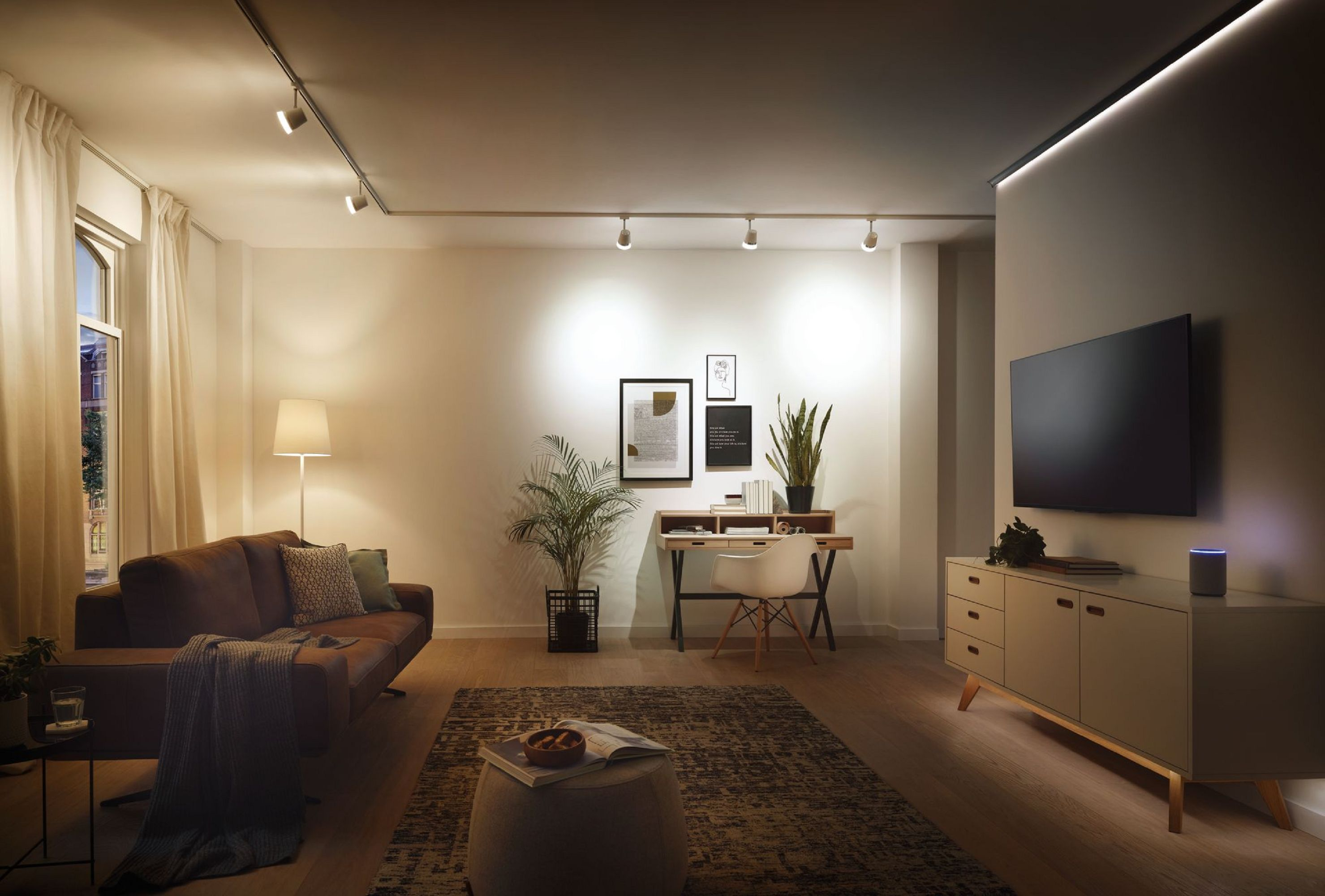 Ein modernes Wohnzimmer mit braunem Sofa, Schreibtisch, Pflanzen und Fernseher. Schienenbeleuchtung beleuchtet den Raum.