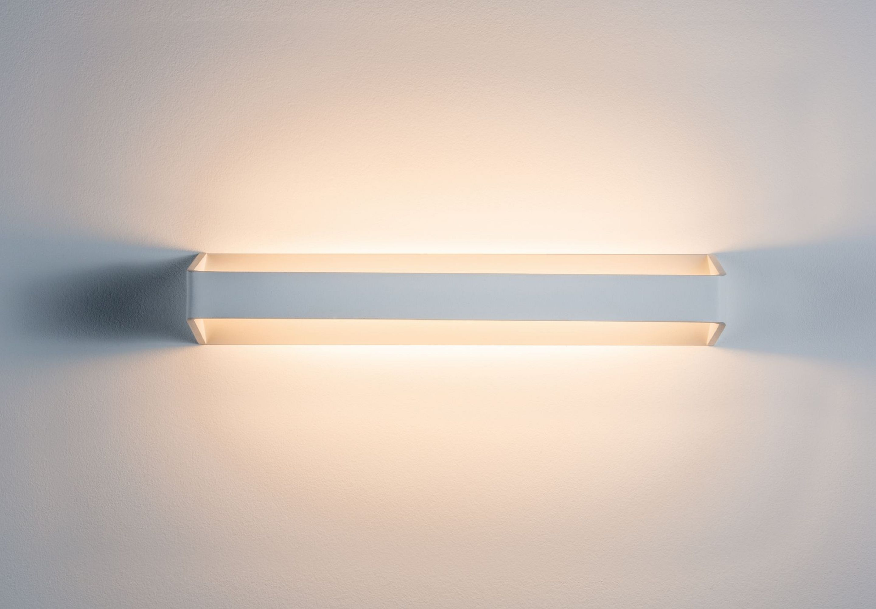 Eine weiße, rechteckige Wandleuchte ist an einer weißen Wand montiert. Die Lampe strahlt nach oben und unten Licht aus.