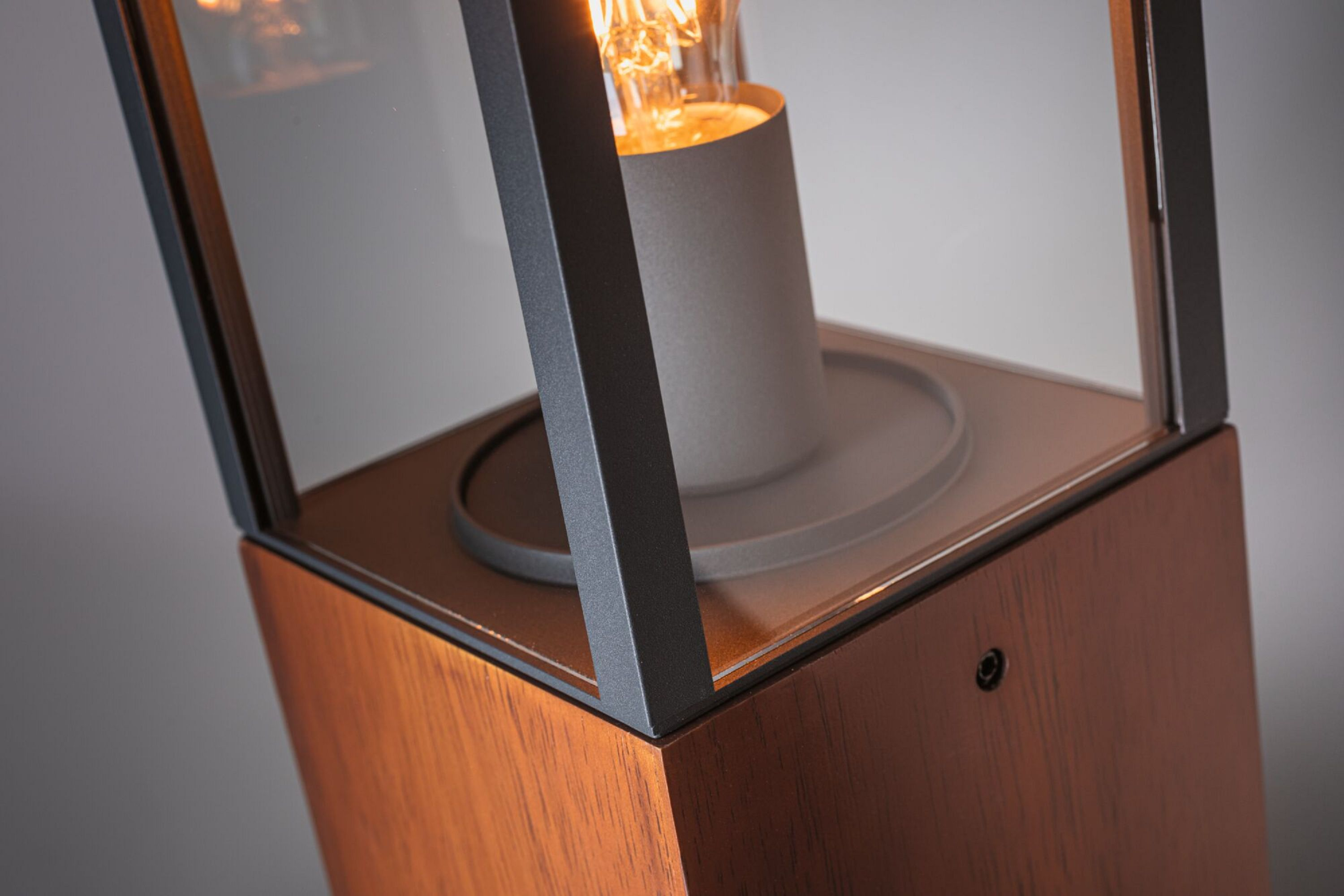 Zbliżenie lampy. Ma drewnianą podstawę, szklane boki, czarną ramę i świecącą żarówkę.