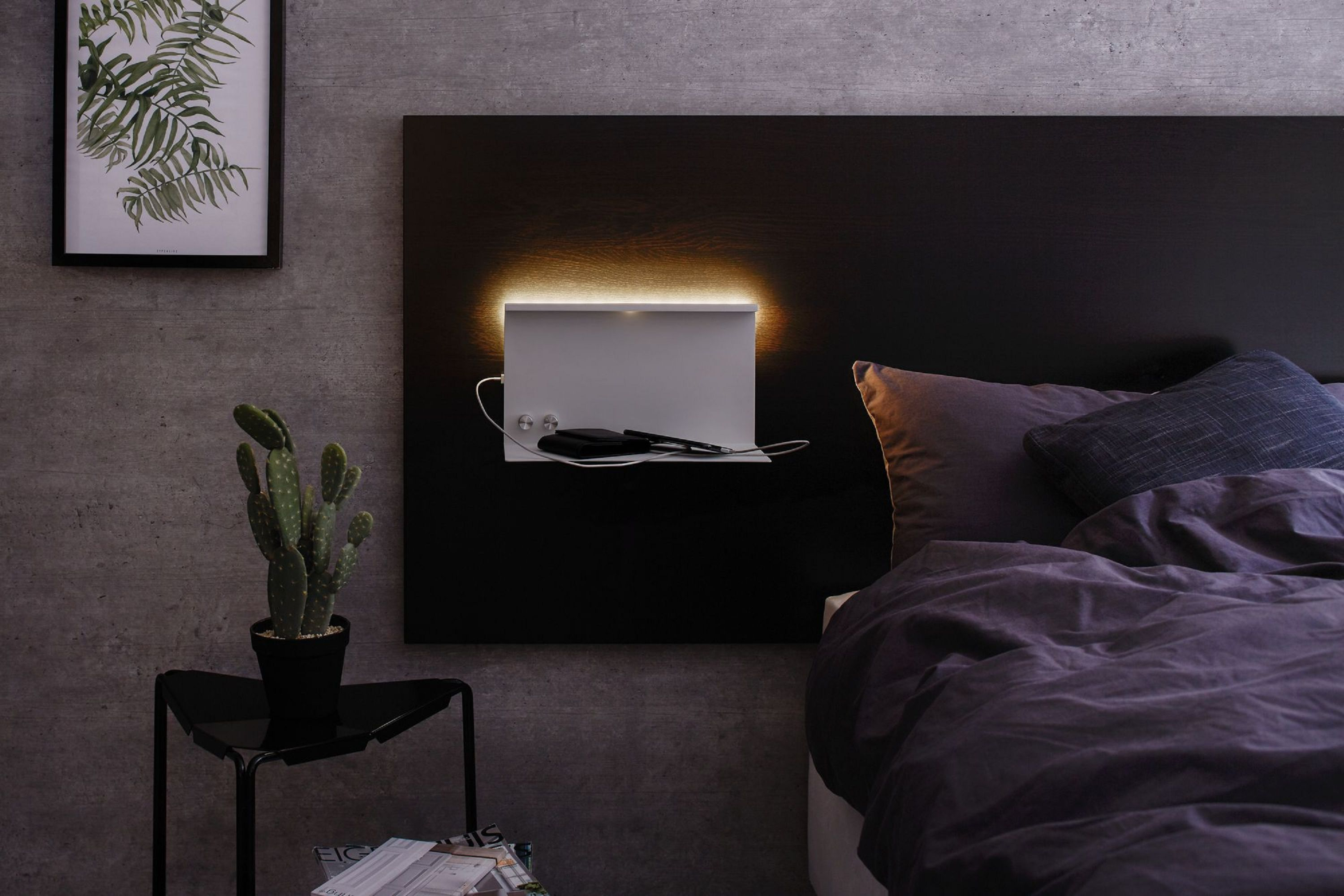 Schlafzimmer mit Bett, Wandregal mit Gegenständen und einem gerahmten Farnprint. Ein Kaktus steht auf einem Beistelltisch.