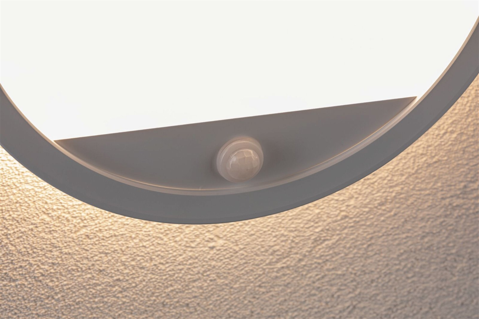 Eine weiße, kreisförmige Deckenleuchte mit einem Bewegungssensor. Das Licht ist teilweise beleuchtet. Der Hintergrund ist eine strukturierte, beige Wand.