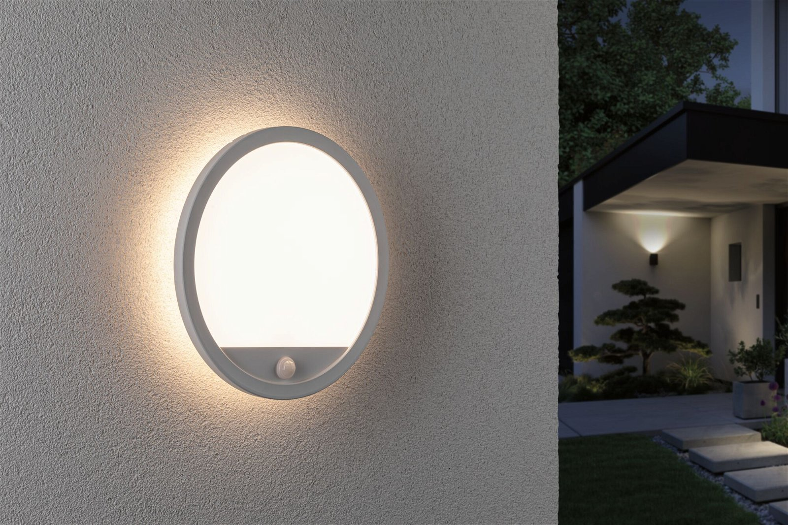 Eine runde Wandleuchte leuchtet an einer strukturierten Wand. Im Hintergrund ein Haus mit beleuchteter Lampe und kleinem Baum.