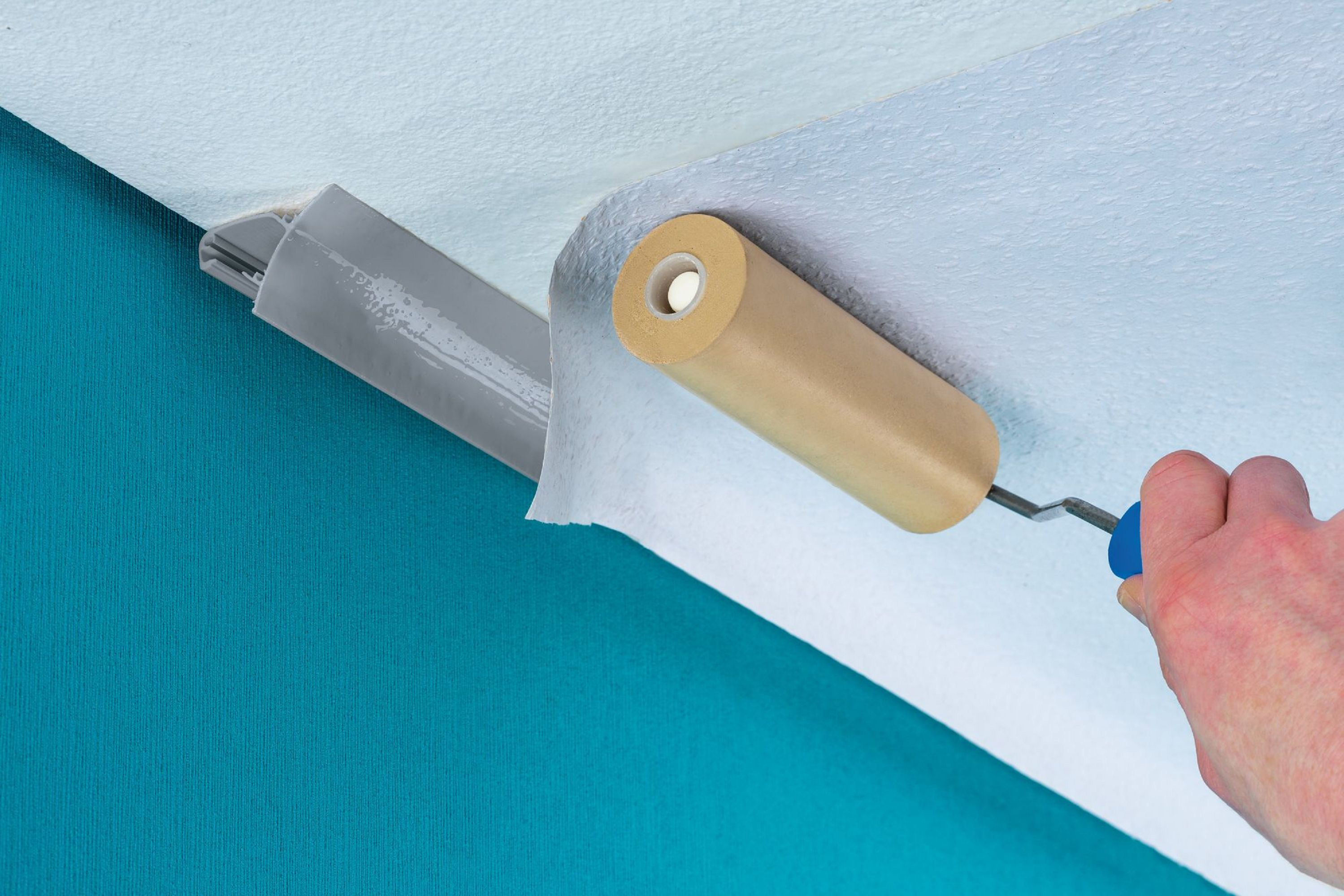 Ręka używa wałka do nakładania tapety na sufit. Turkusowa ściana po lewej. Wałek jest beżowy i szary.
