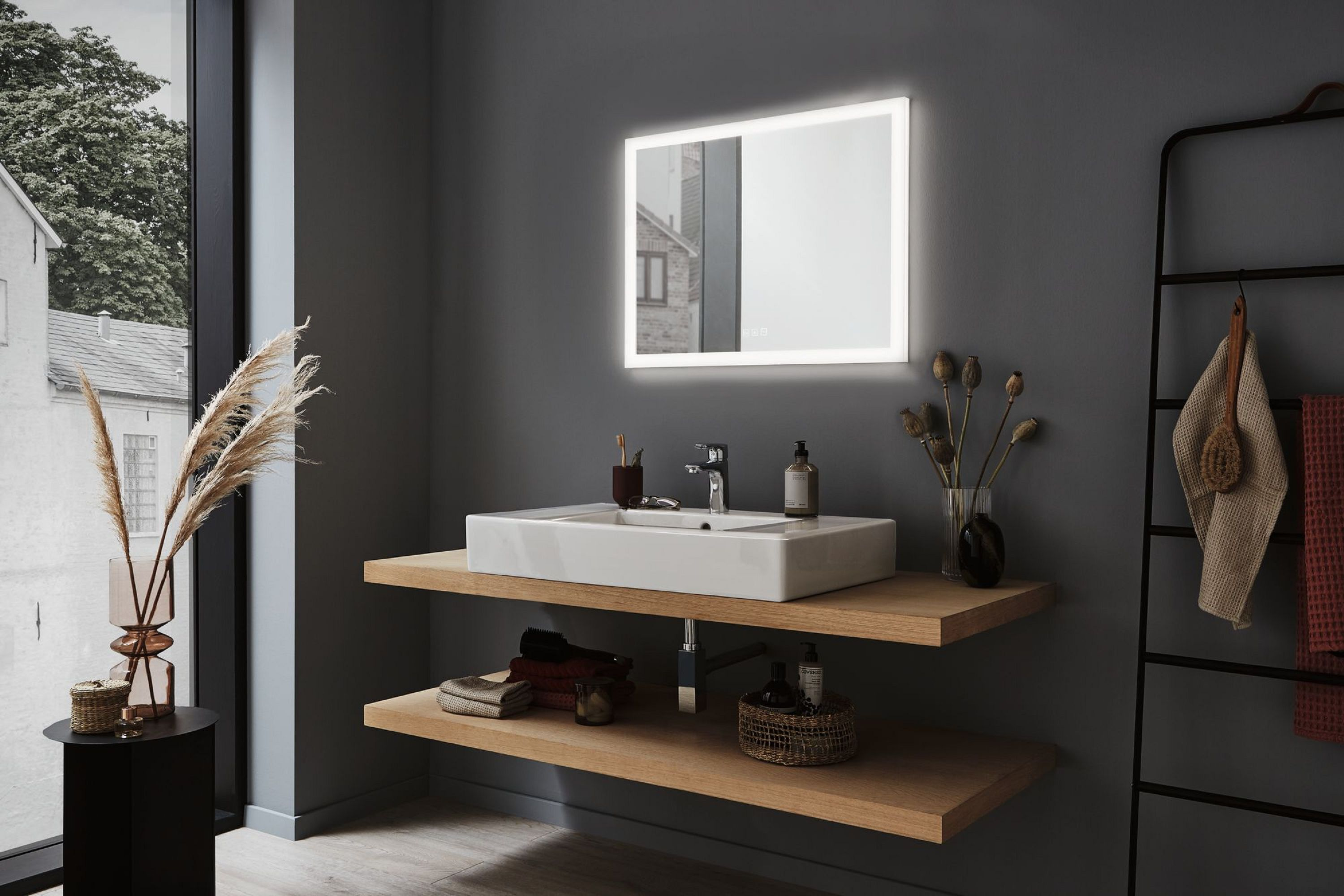 Badezimmer mit rechteckigem Spiegel, Waschbecken und Holzregalen. Ein Fenster zeigt ein Gebäude. Dekorative Gegenstände sind vorhanden.