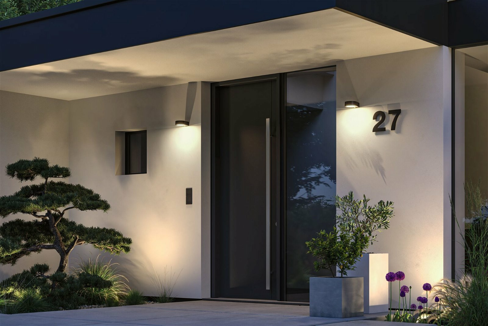 Nowoczesne wejście do domu w nocy z czarnymi drzwiami, numer 27, roślinami i światłami. Drzwi są uchylone.