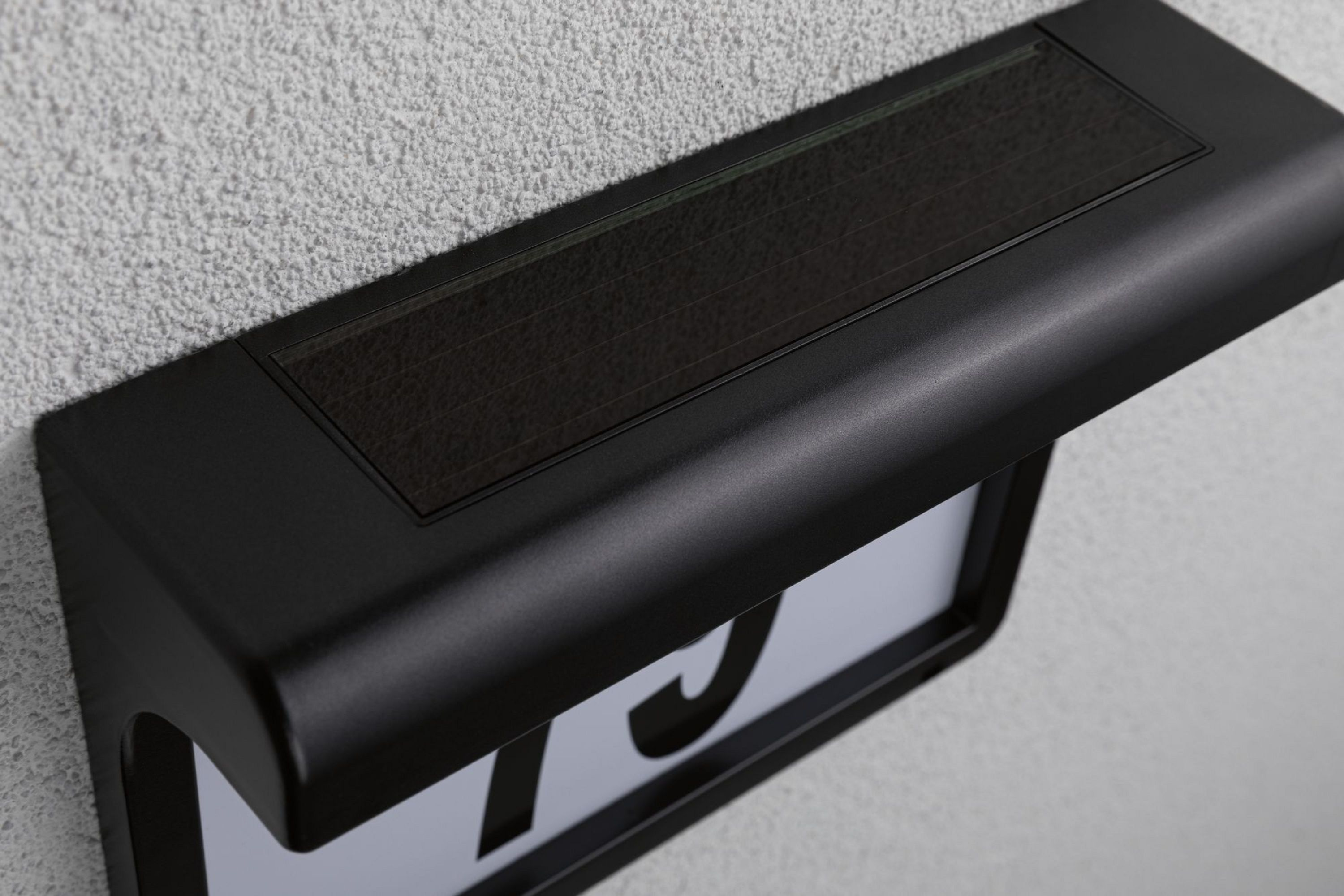 Czarny, zasilany energią słoneczną szyld adresowy jest zamontowany na teksturowanej białej ścianie. Szyld wyświetla numer.