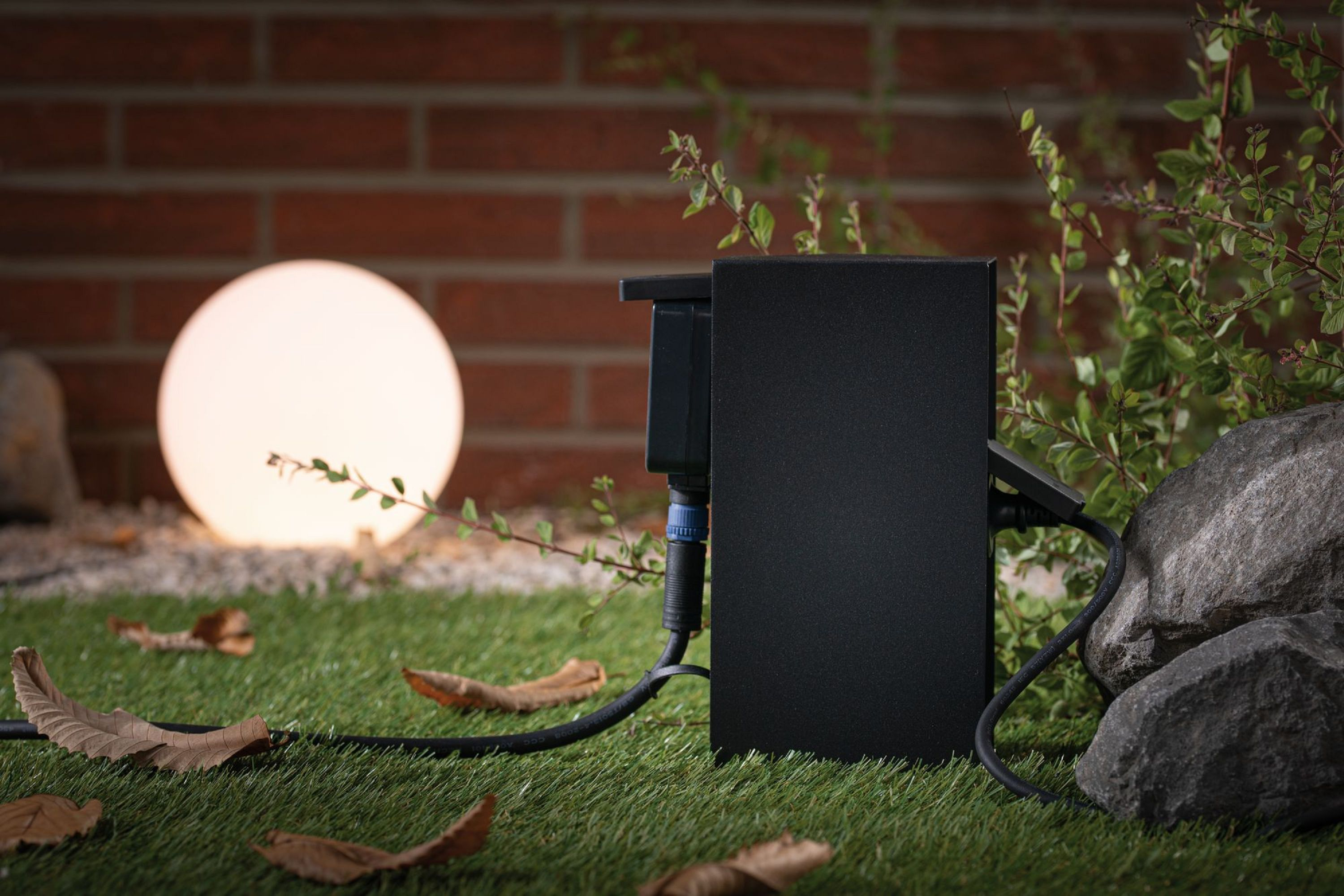 Czarna skrzynka elektryczna z kablem na trawie, świecąca lampa kulista i ceglany mur w tle.