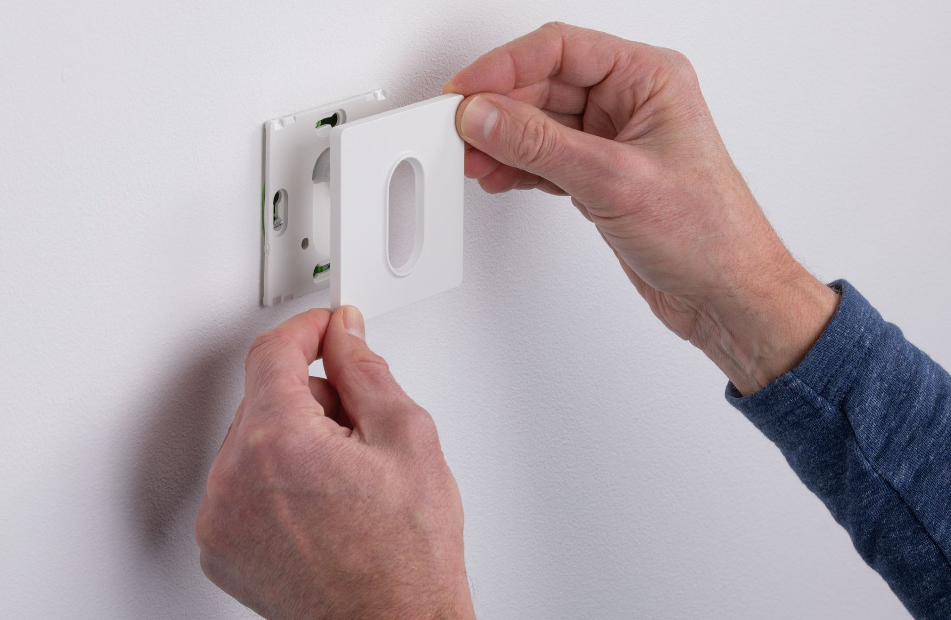 Hände installieren eine weiße Abdeckplatte an einer weißen Wand. Die Platte hat eine ovale Öffnung.