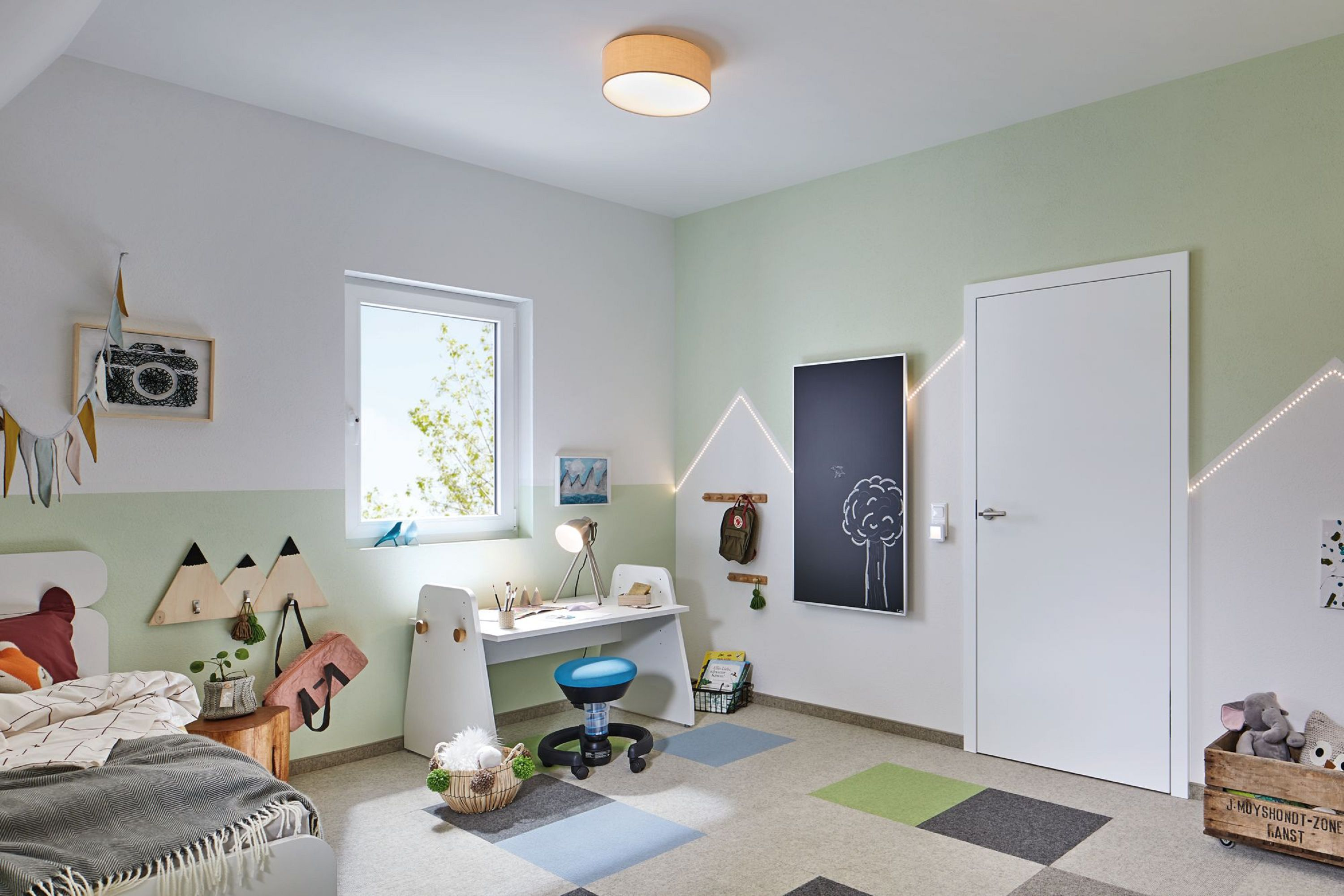 Kinderzimmer mit Schreibtisch, Bett und Tafel. Das Zimmer ist mit einem Kamera-Bild und dreieckigen Haken dekoriert.