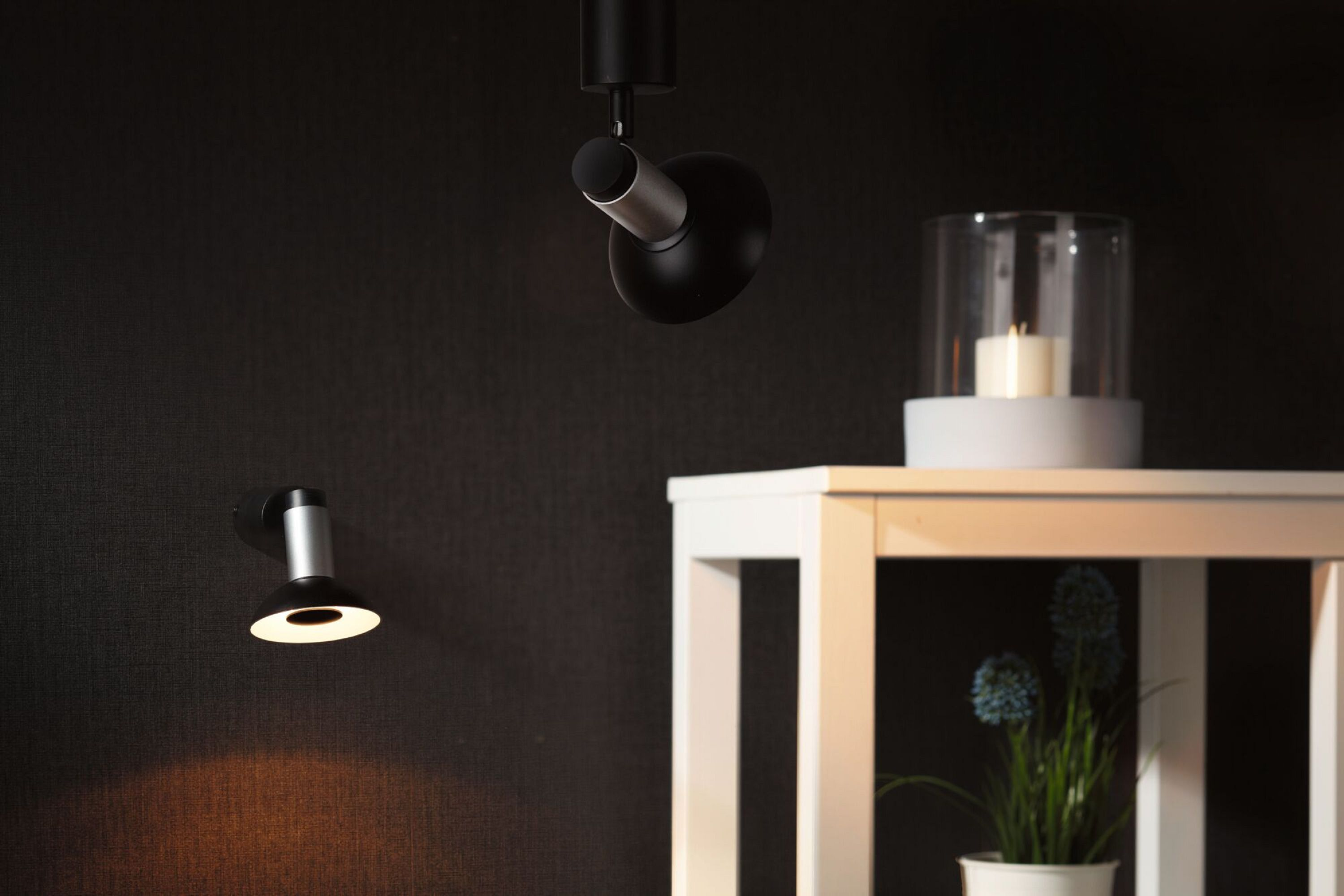 Schwarze und silberne Lampen, weißer Tisch mit Kerze und Pflanze mit blauen Blumen. Dunkler Hintergrund.