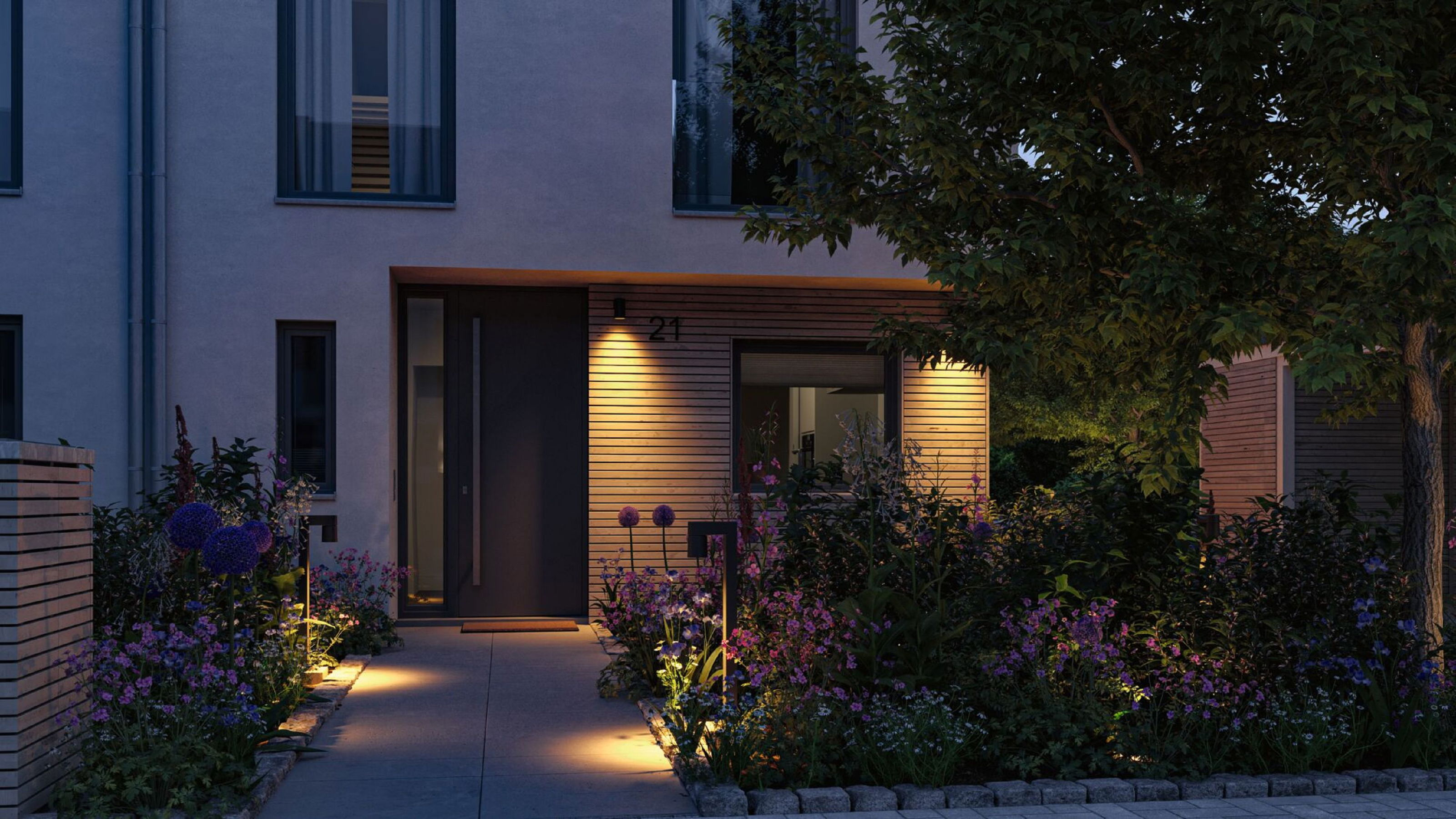 Zewnętrze nowoczesnego domu o zmierzchu, z ogrodem kwiatowym. Drzwi wejściowe mają numer 21. Ciepłe światła oświetlają scenę.