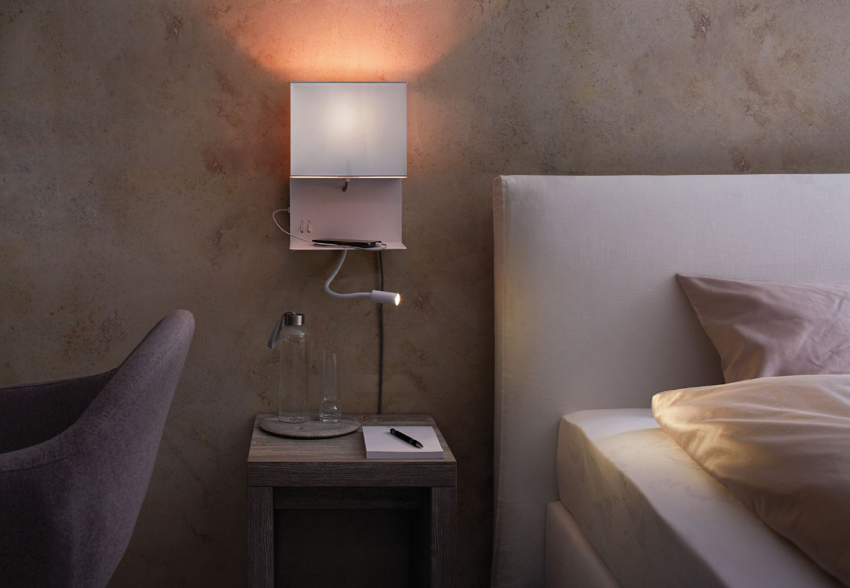Eine Nachttischszene mit Lampe, Telefon und Wasserflasche auf einem Nachttisch neben einem Bett.