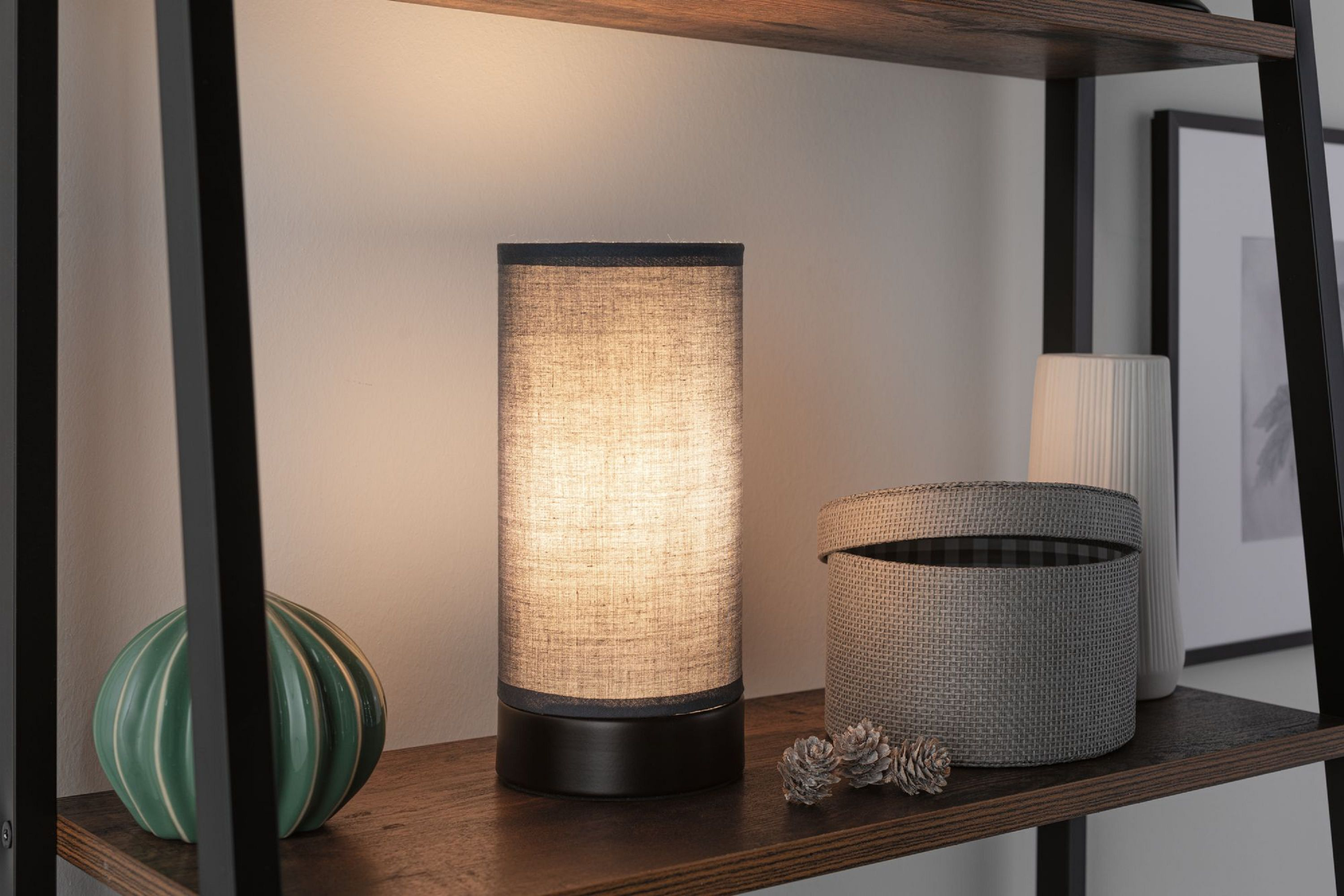 Una lampada, un cestino, un vaso e un oggetto decorativo sono su uno scaffale. Lo sfondo è una parete bianca e un telaio in metallo nero.