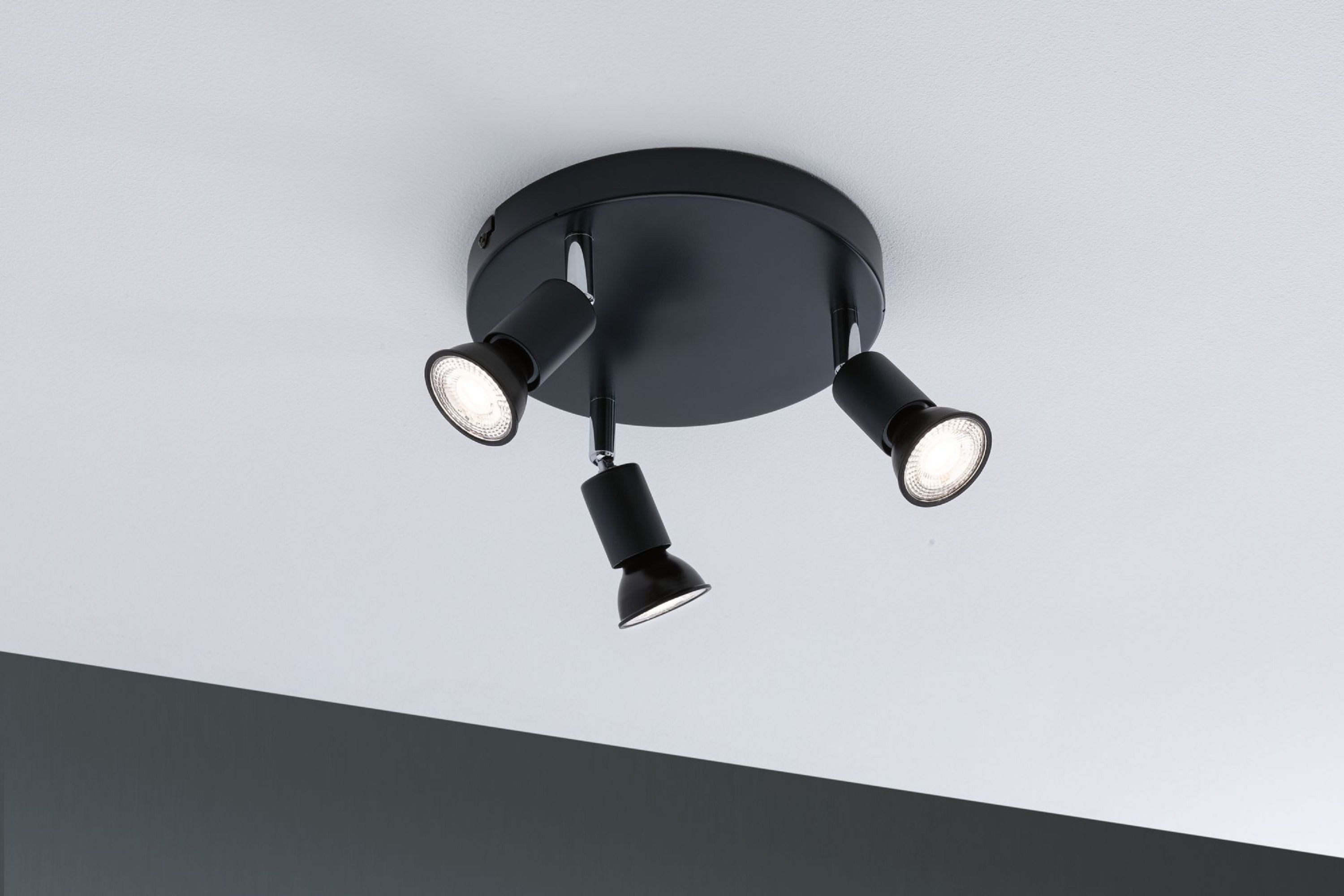 Schwarzer Deckenstrahler mit drei verstellbaren Lichtern vor weißer Decke. Die Lichter sind an.