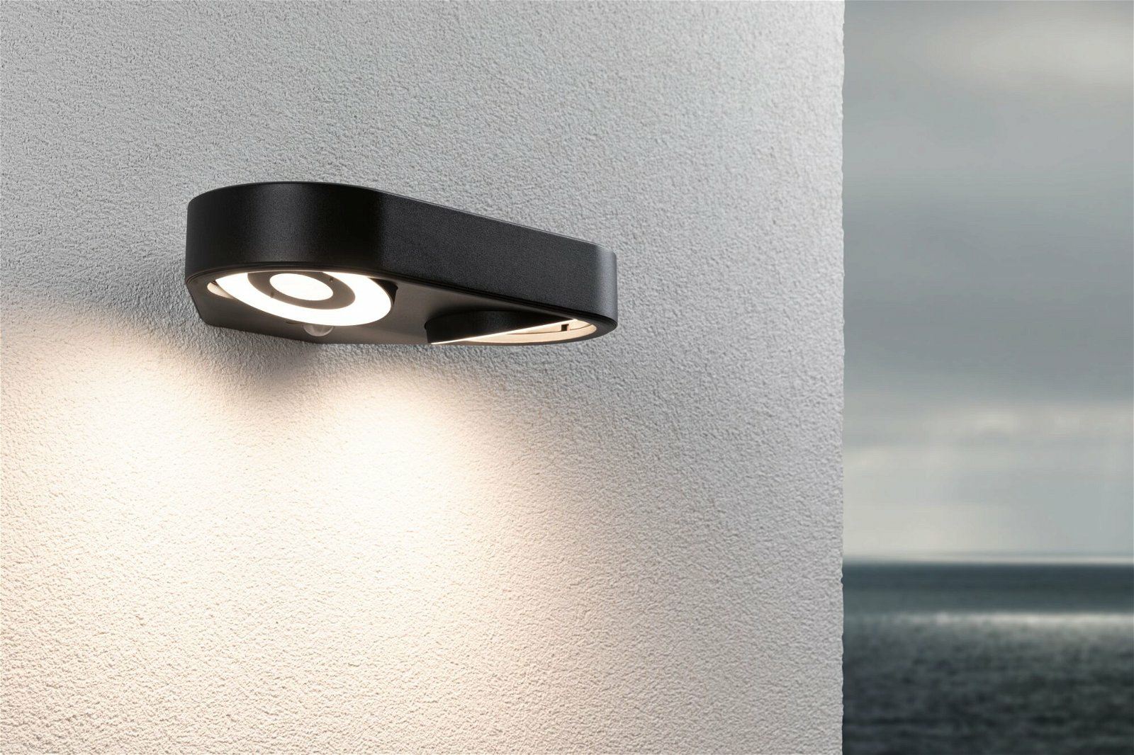 Czarna zewnętrzna lampa ścienna z jasnym światłem na teksturowanej białej ścianie. Tło to rozmyte morze i niebo.