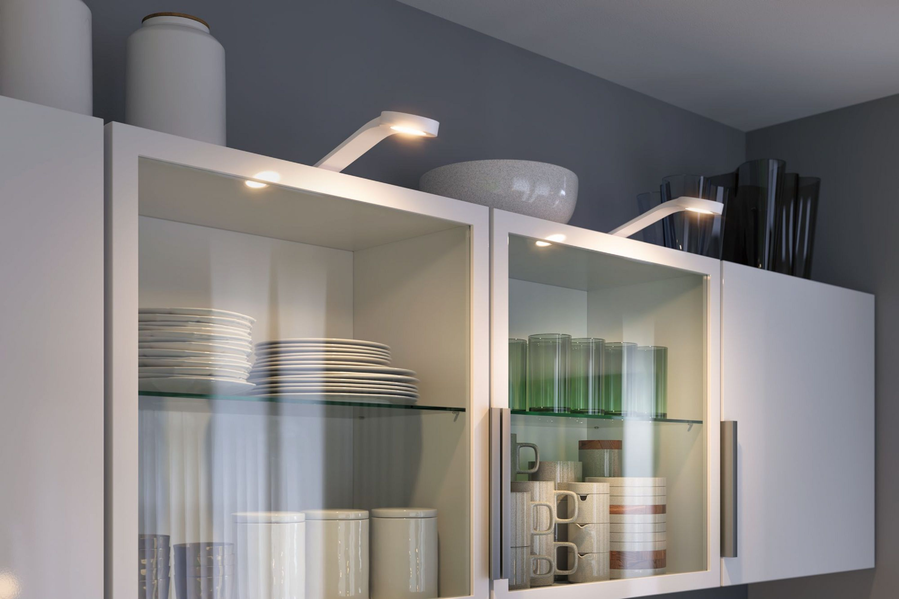 Mobile da cucina con ante in vetro. All'interno, piatti, bicchieri e tazze sono visibili. Due lampade bianche illuminano.