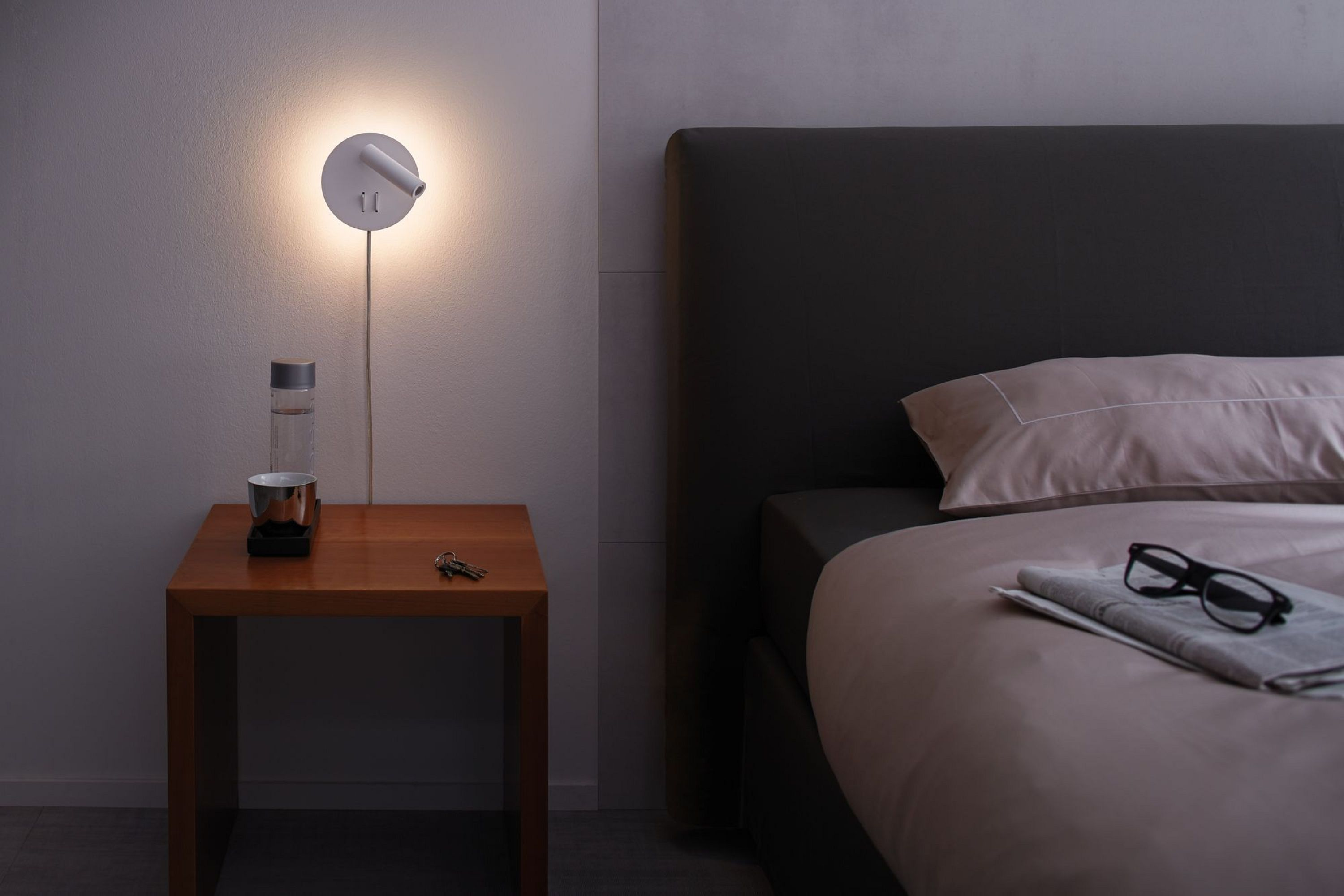 Schlafzimmer mit Bett, Nachttisch, Lampe und Wasserflasche. Brille und Zeitung liegen auf dem Bett.