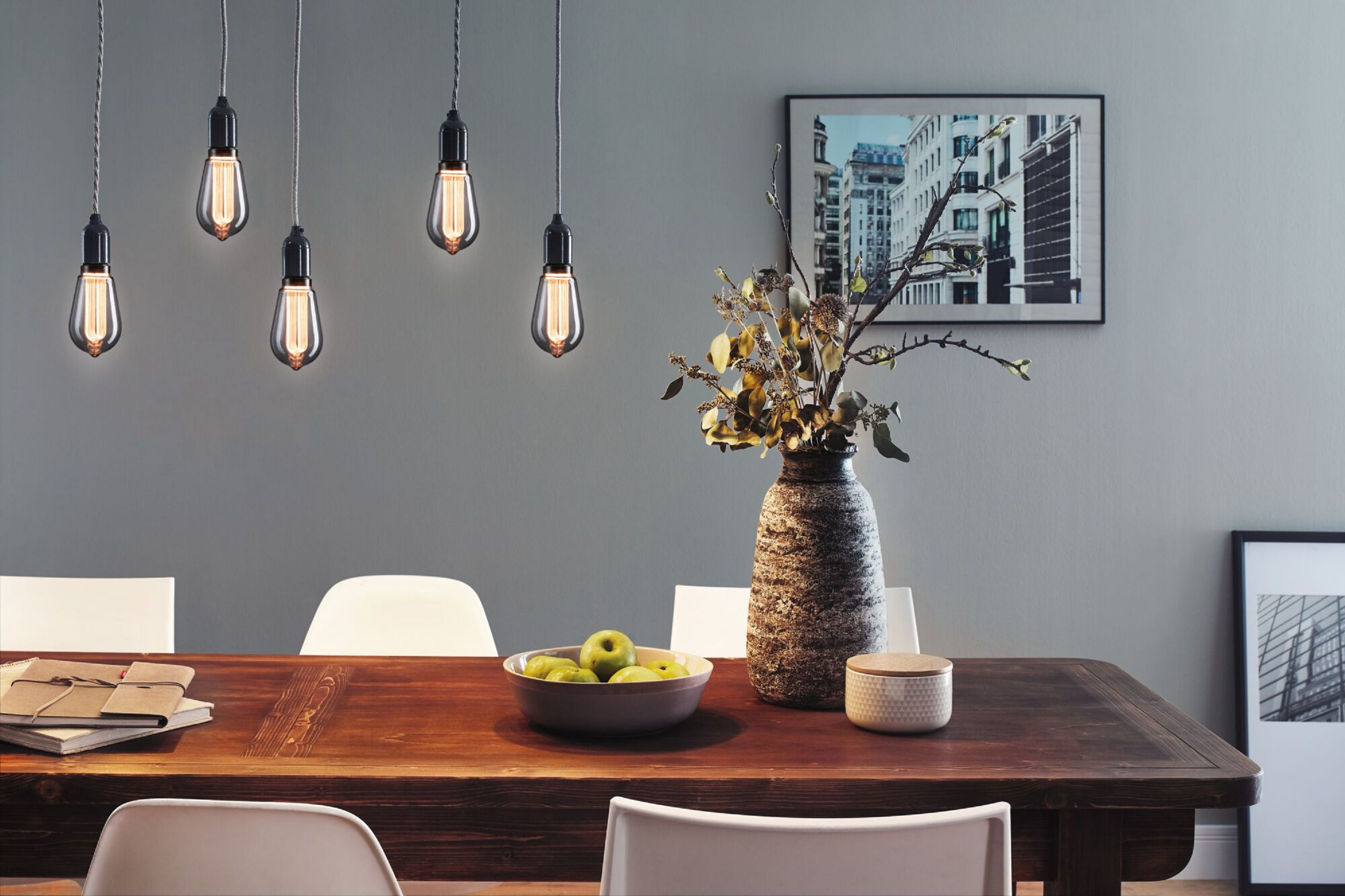 Esszimmer mit Hängelampen, Holztisch, Äpfeln in einer Schüssel, Vase mit Blumen und gerahmten Bildern.