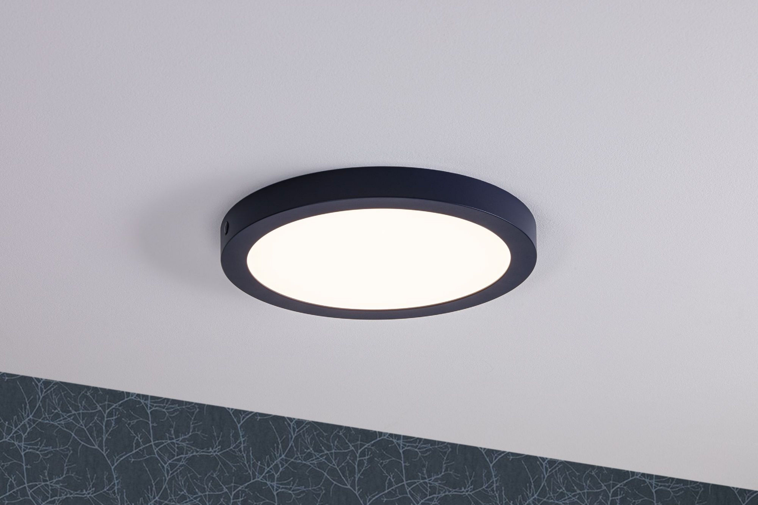 Un plafonnier rond noir avec un centre blanc brillant est monté sur un plafond blanc. Du papier peint est visible en dessous.
