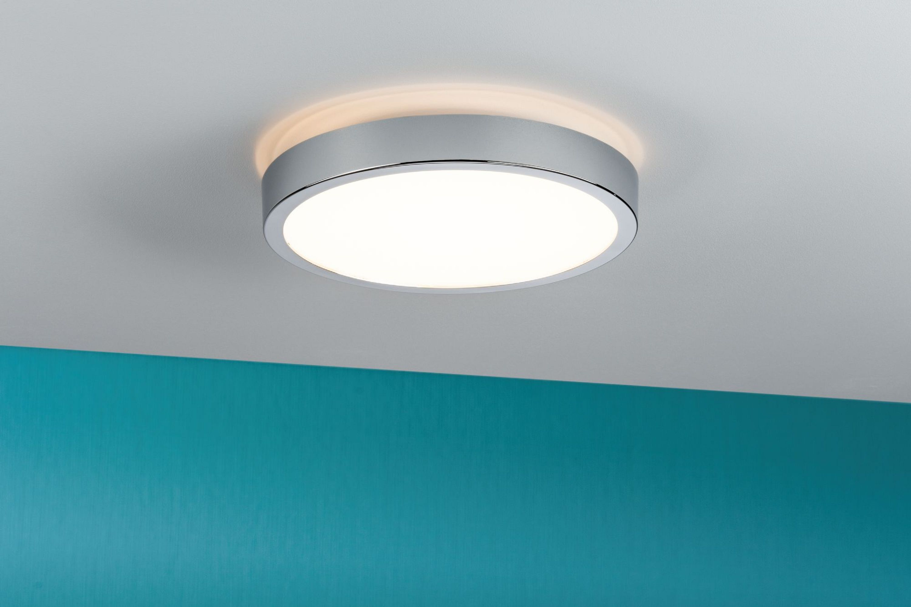 Un plafonnier rond et argenté est centré contre un plafond blanc. Un mur turquoise est en dessous. La lumière est allumée.