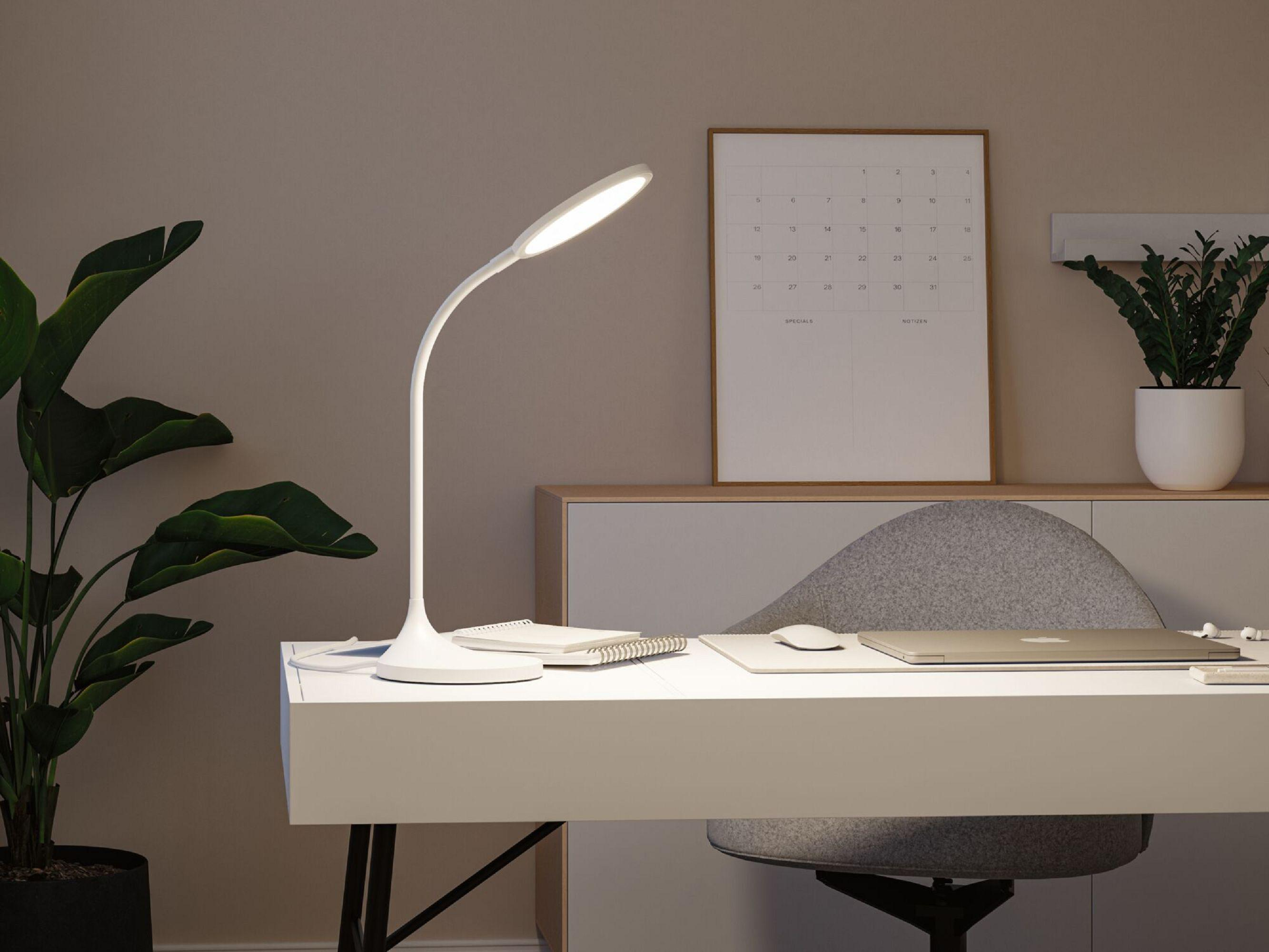 Na zdjęciu biała lampa biurkowa, biurko, kalendarz, laptop, rośliny i krzesło.