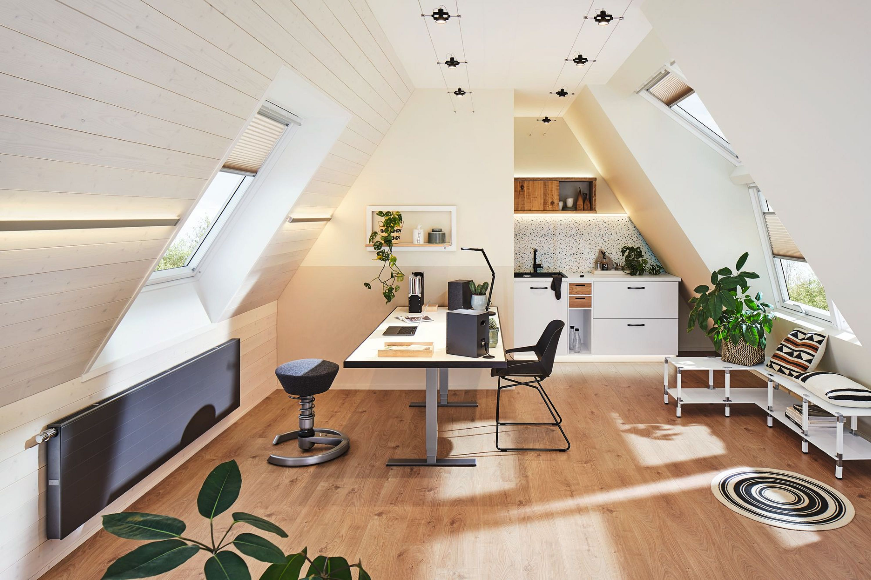 Poddasze z biurkiem, kuchnią i częścią wypoczynkową. Widoczne są okna dachowe, drewniane podłogi i ciemnoszary grzejnik.