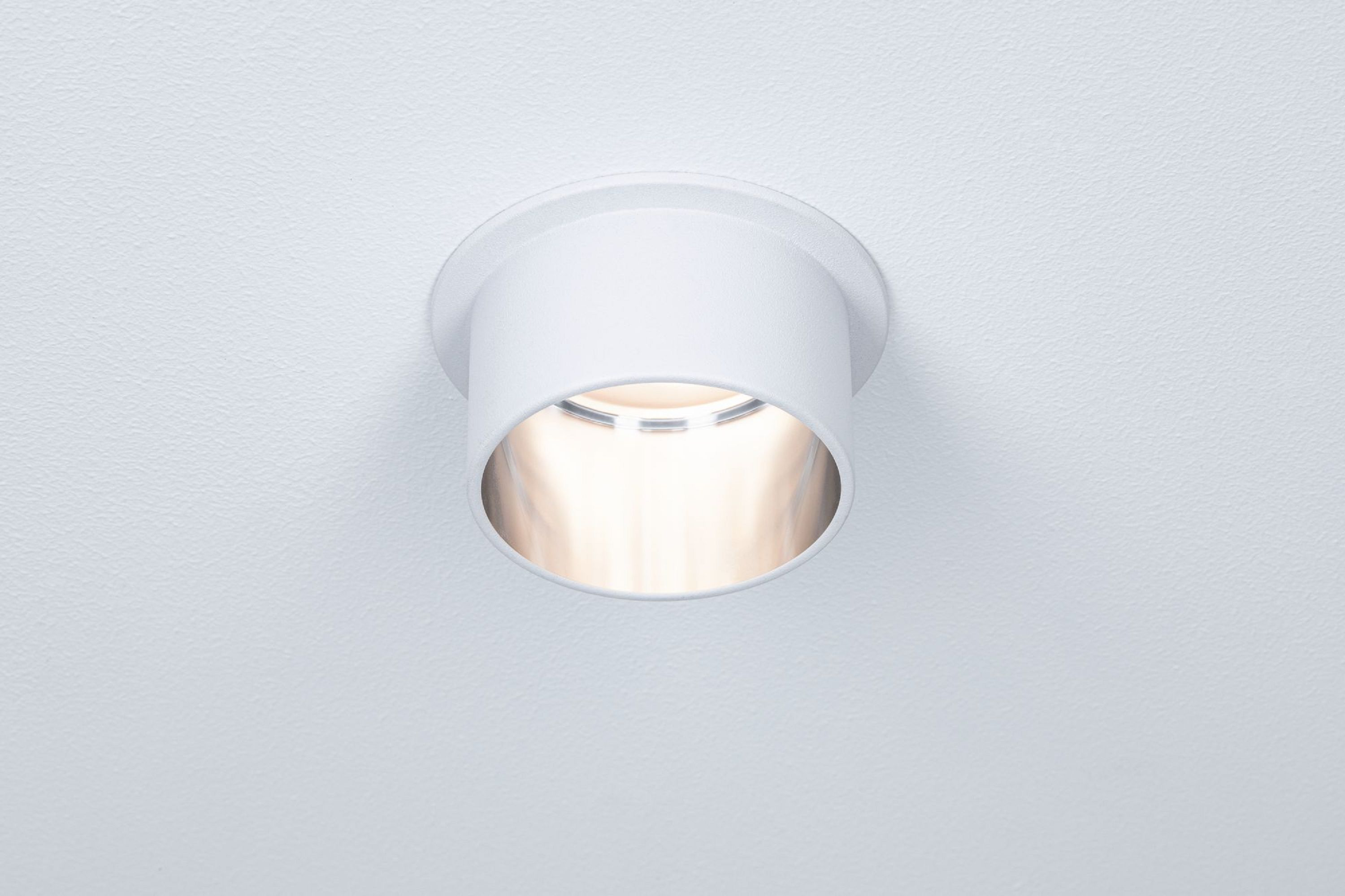 Eine weiße Einbauleuchte mit zylindrischer Form. Das Licht ist an und beleuchtet die Öffnung vor einem weißen Hintergrund.
