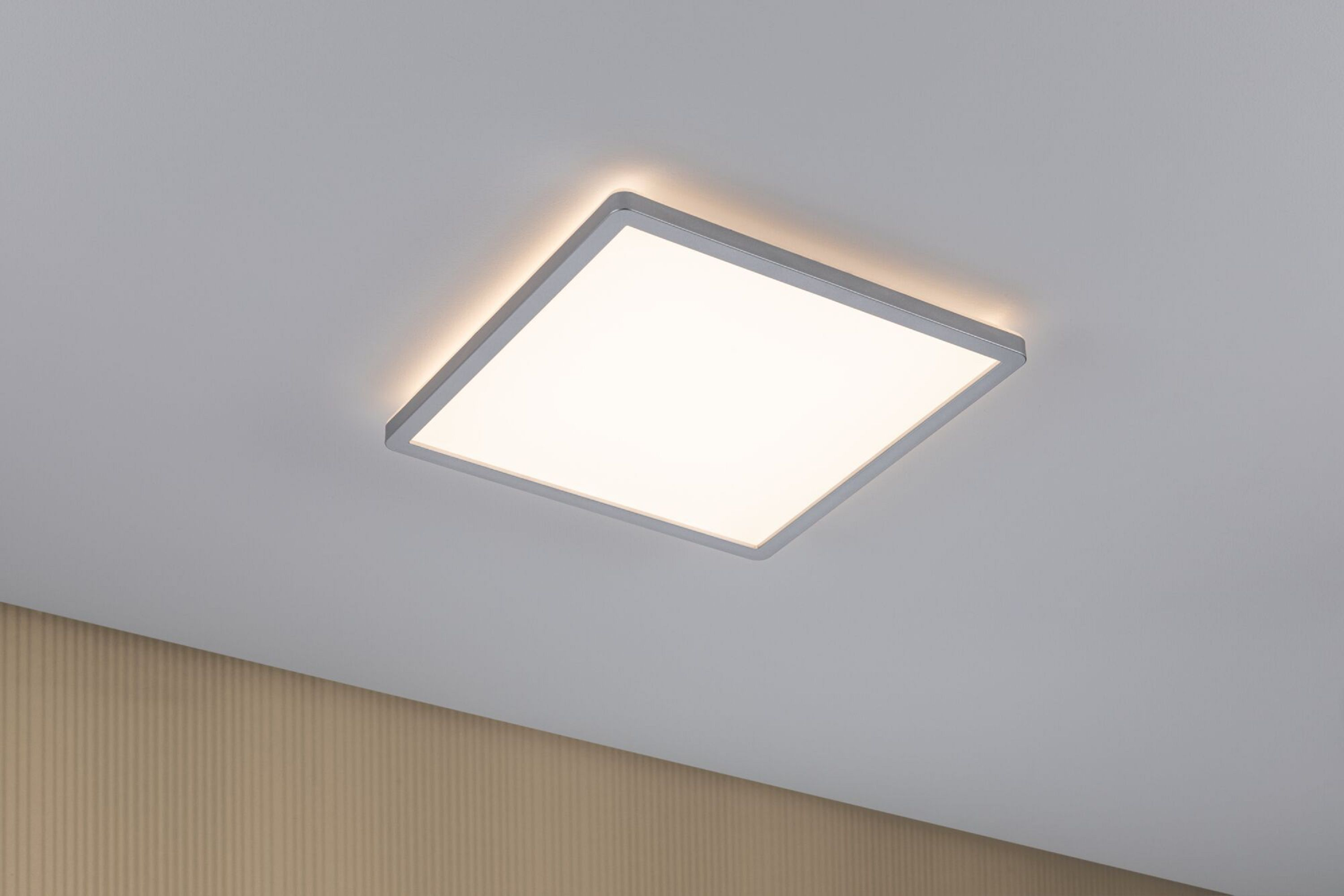 Kwadratowa, wpuszczana lampa sufitowa ze srebrną ramą oświetla pomieszczenie. Beżowa ściana na dole po lewej.
