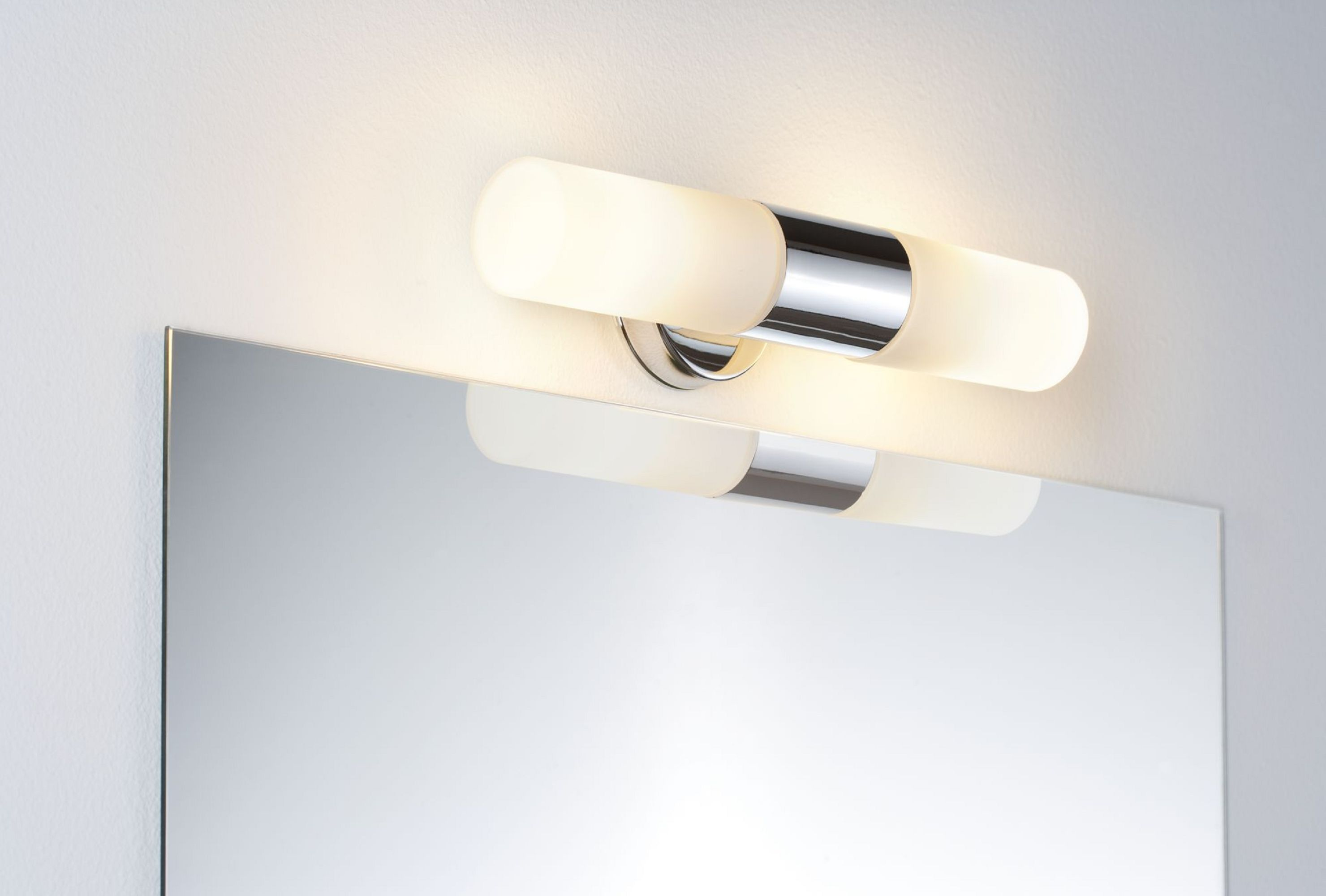 Uno specchio da bagno con un'apparecchio di illuminazione sopra. La luce è cromata e in vetro smerigliato. Il muro è bianco.