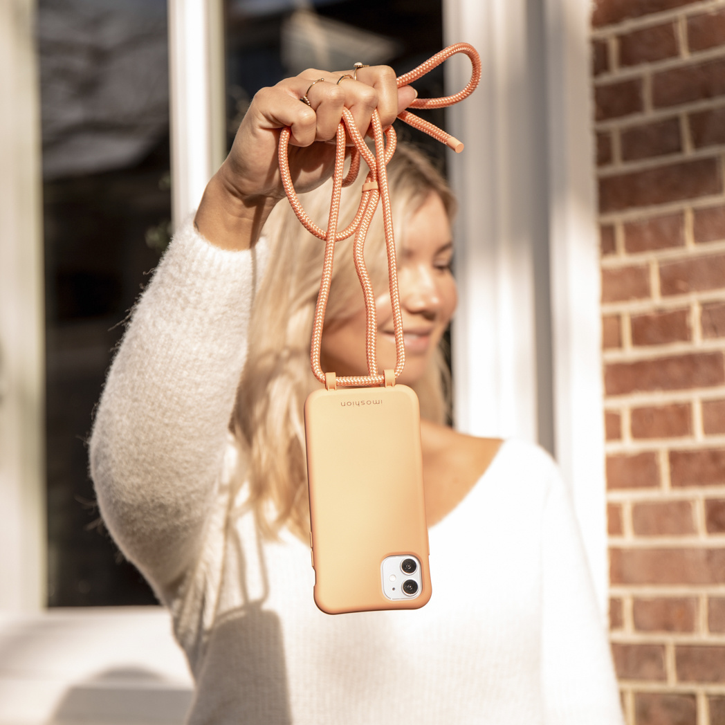 Een vrouw houdt een perzik-kleurige telefoon omhoog met een bijpassend koord. Ze draagt een witte trui, met een bakstenen muur en een raam op de achtergrond.