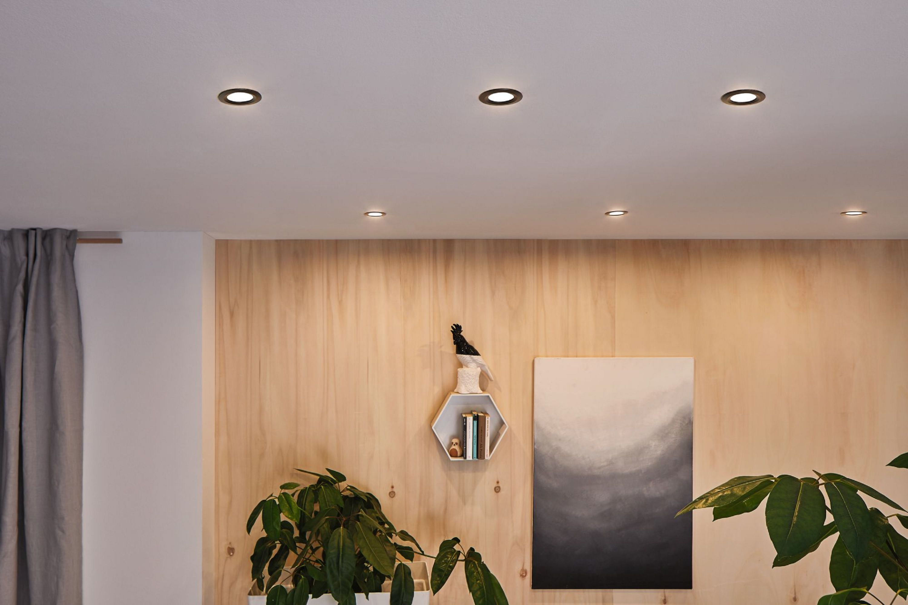 Innenansicht mit Deckenleuchten, Holzwand, Kunst, Pflanzen und Vorhängen. Ein moderner, gut beleuchteter Wohnraum.