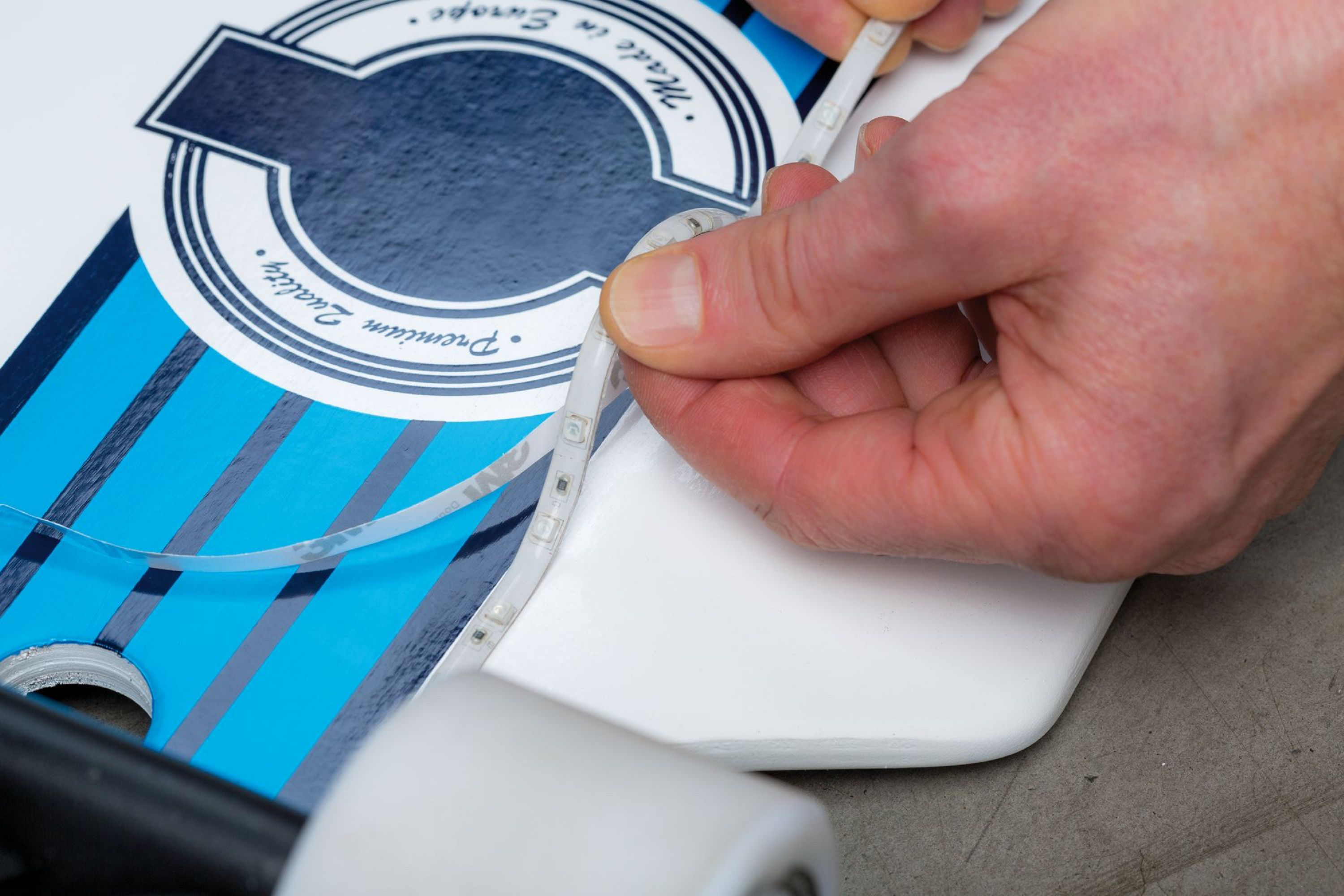 Eine Hand zieht einen LED-Streifen von einem Skateboard ab. Das Board ist weiß und blau, mit einem Logo.