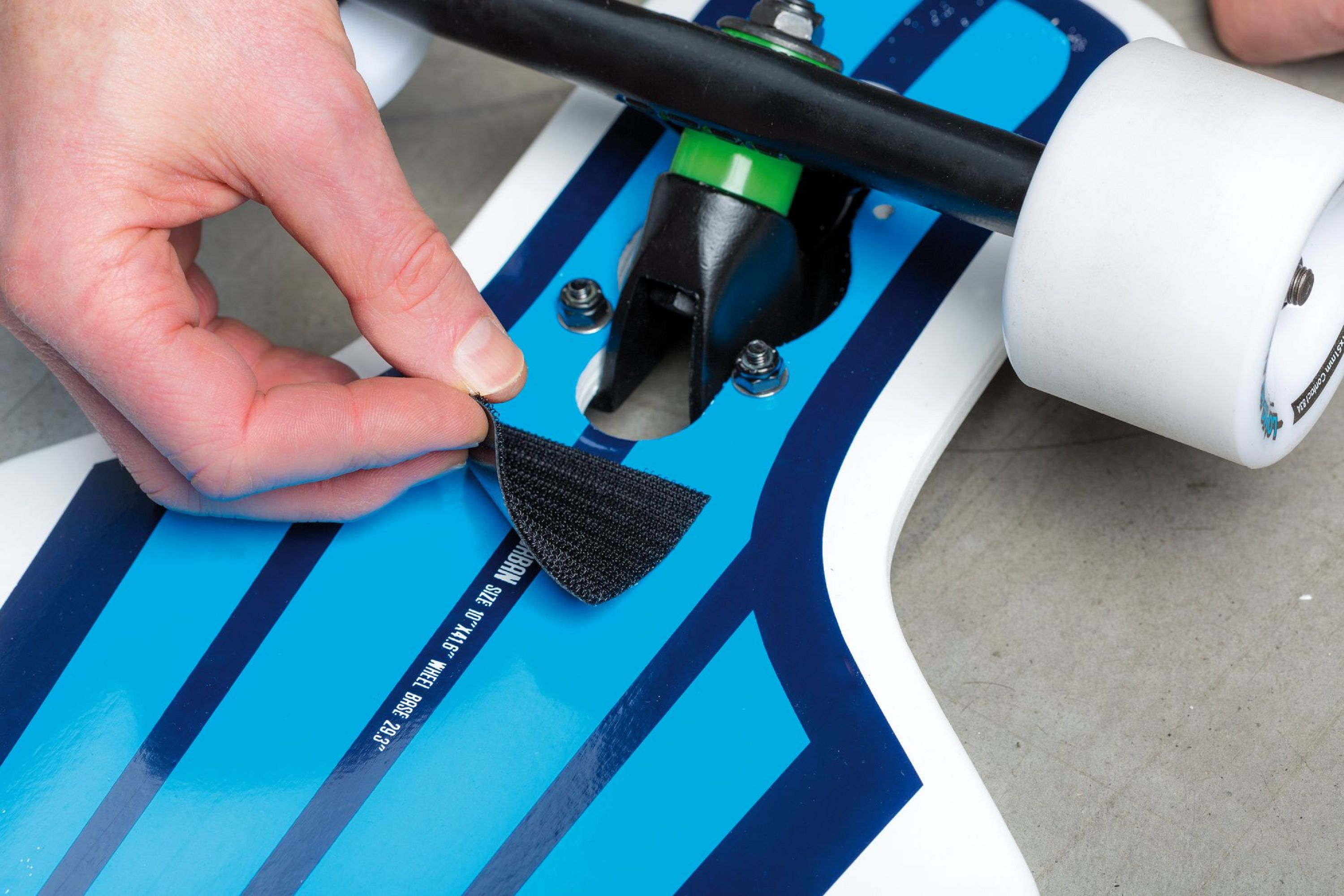 Eine Hand bringt einen schwarzen Klettstreifen an einem blau-weißen Skateboard an. Die Räder und Achsen sind sichtbar.