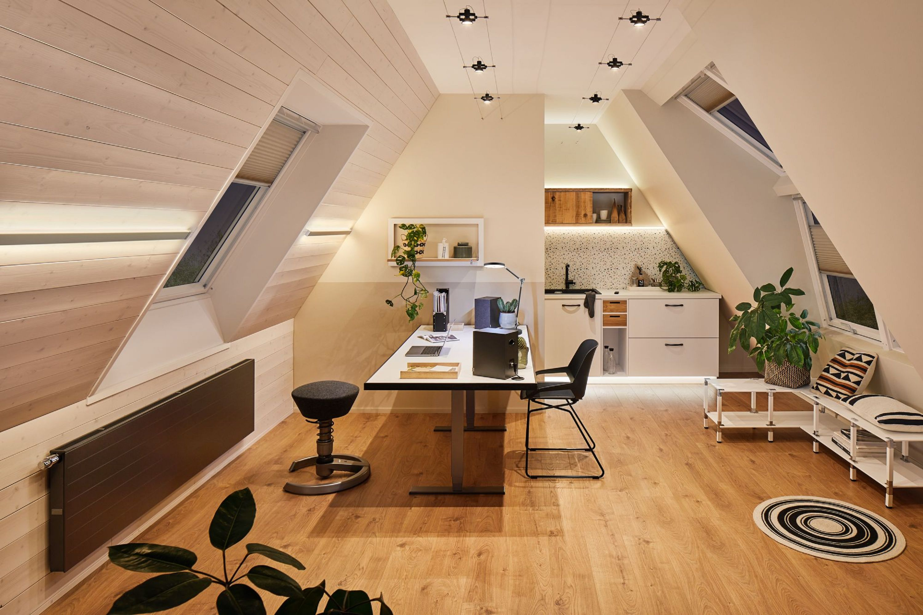 Dachboden mit Schreibtisch, Stuhl und kleiner Küche. Natürliches Licht durch Dachfenster. Holzboden und Wände.