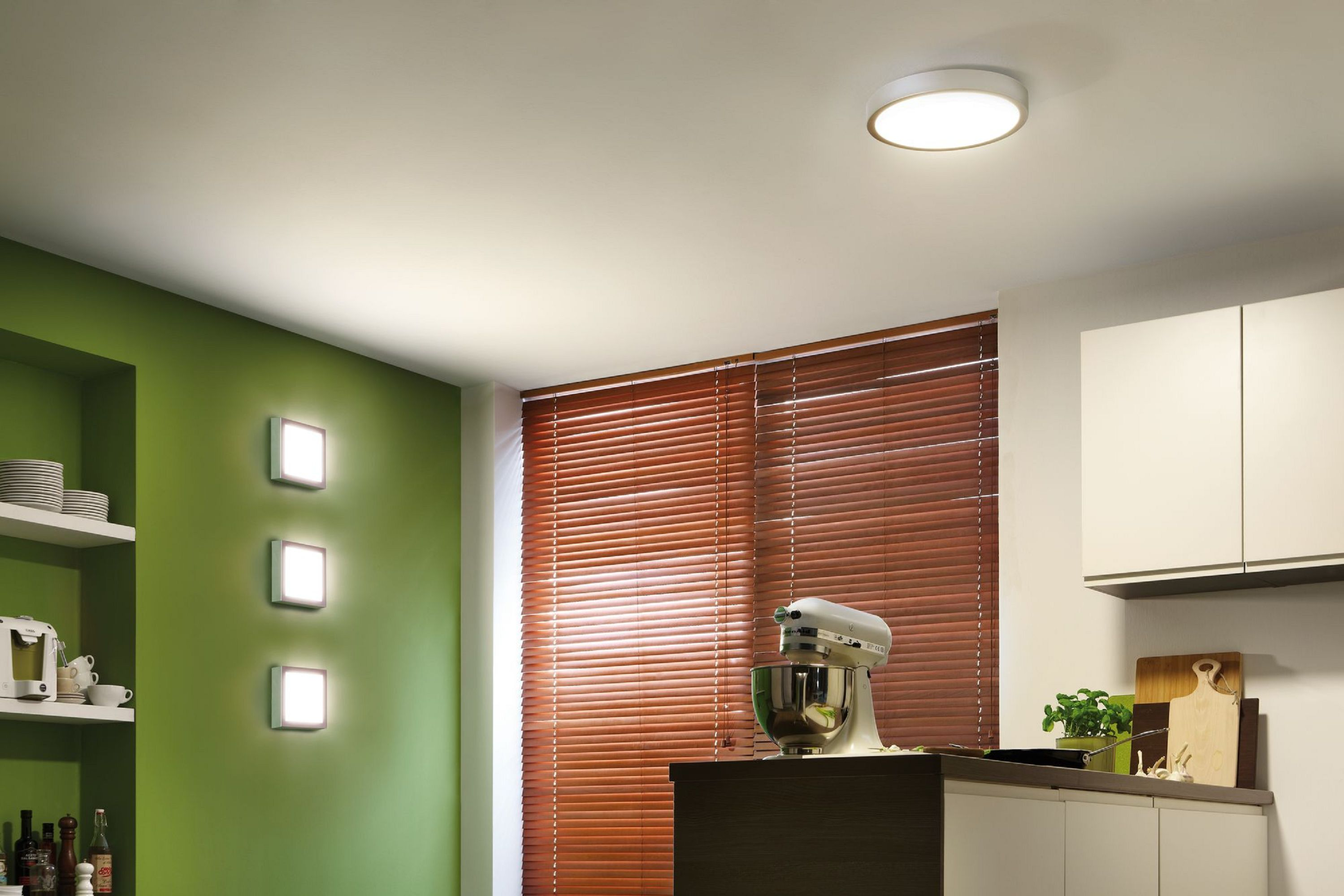 Scena cucina con parete verde, persiane marroni e mixer. La luce del soffitto è accesa. Armadi e scaffali sono visibili.