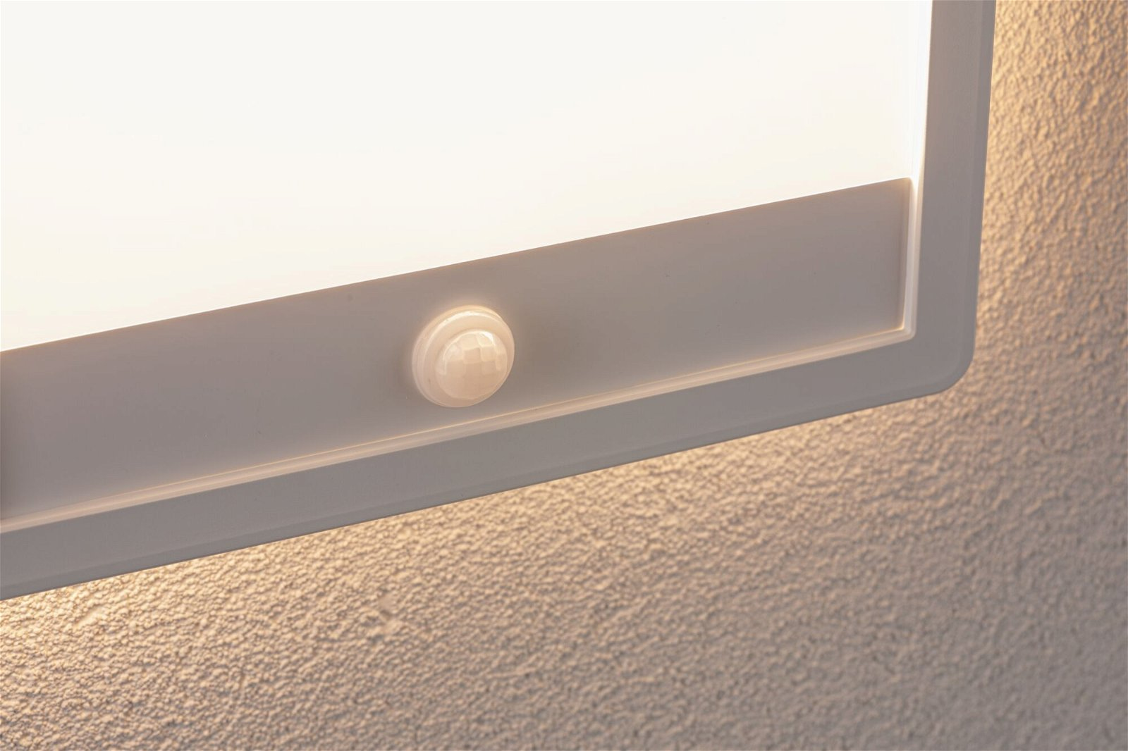 Zbliżenie białej, prostokątnej oprawy oświetleniowej z czujnikiem ruchu. Światło jest jasne, a ściana ma fakturę.