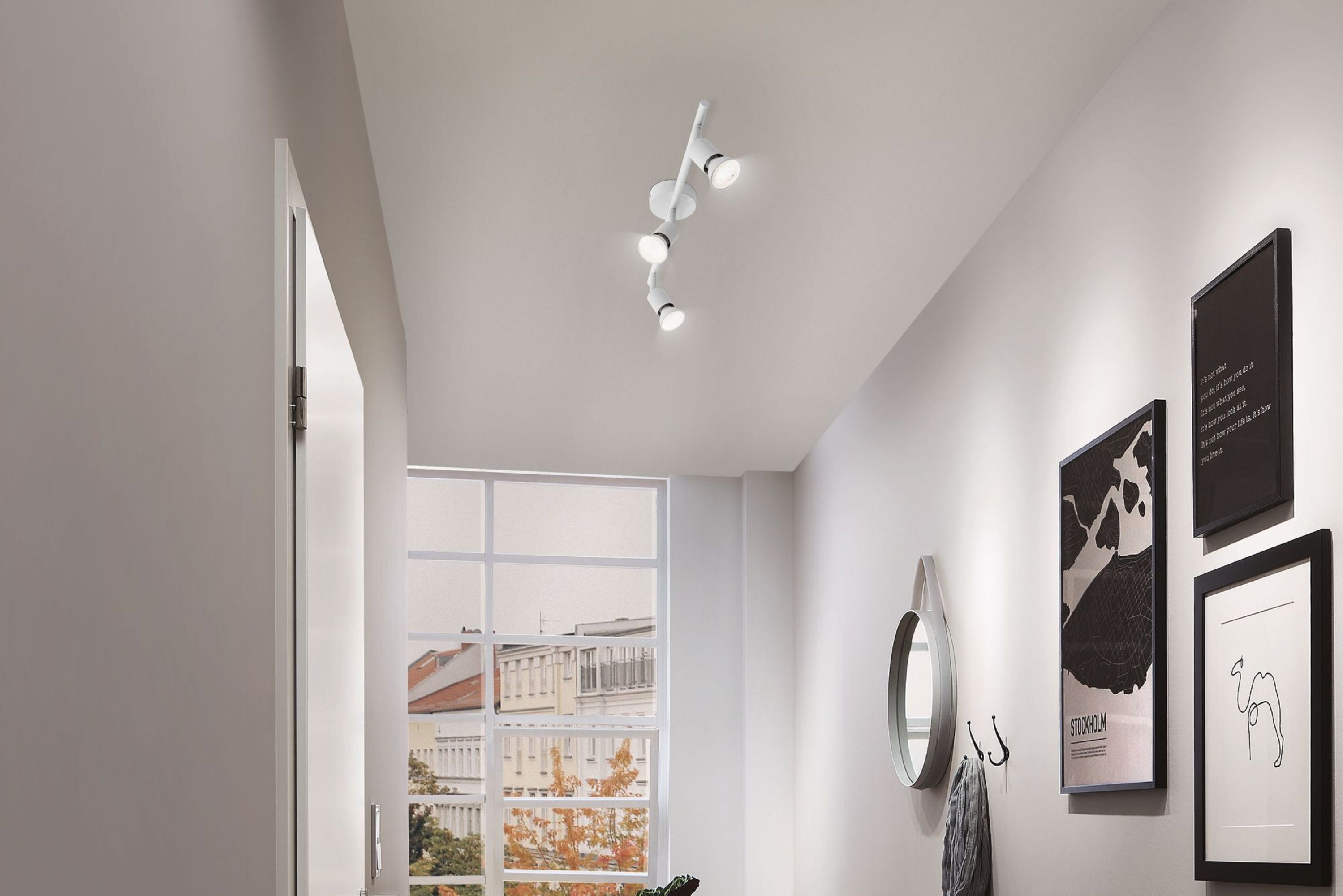 Ein Flur mit weißen Wänden, einer Deckenleuchte und gerahmten Bildern. An der Wand hängen ein Spiegel und Haken, links ist ein Fenster.
