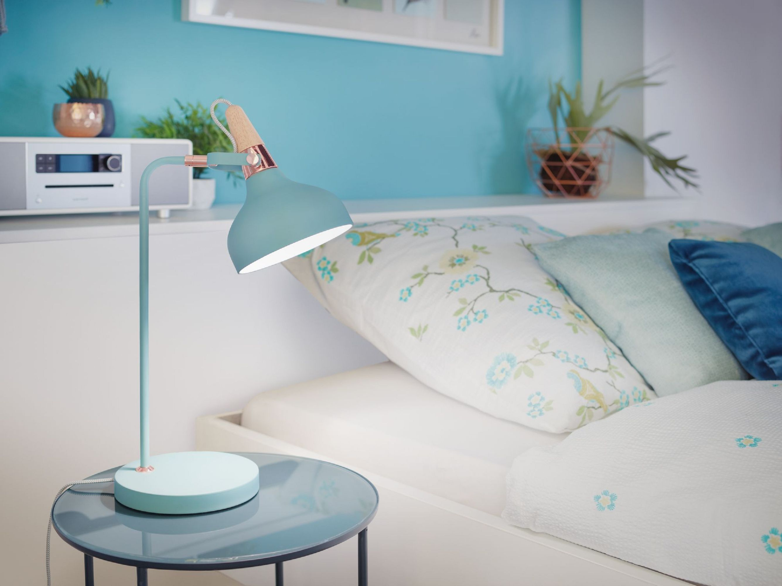 Una lampada turchese illumina un letto con cuscini fantasia. Un comodino, piante e un muro blu completano la scena.