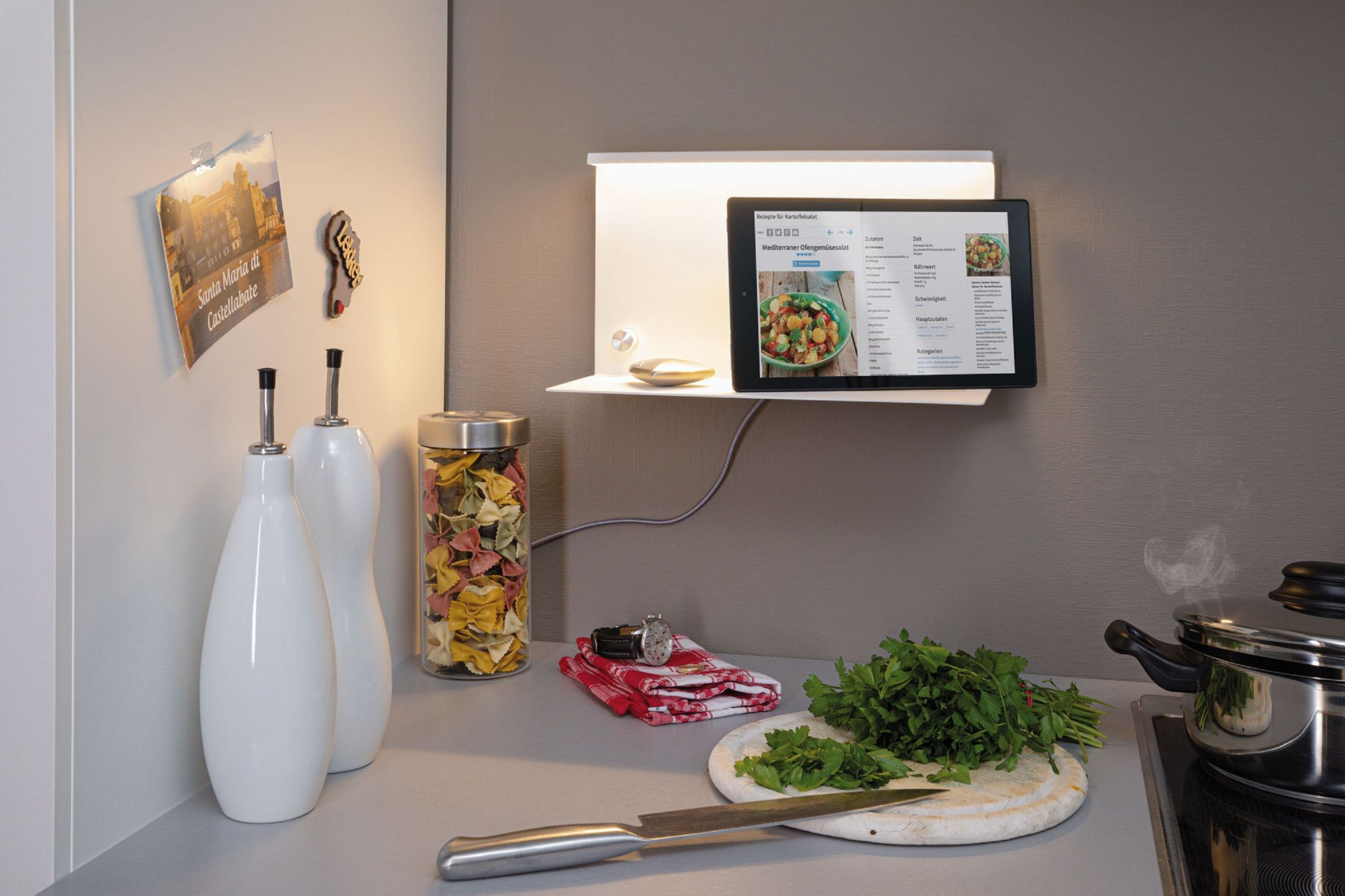 Eine Küchenszene mit einem Tablet, auf dem ein Rezept angezeigt wird. Nudeln, Kräuter und ein Topf stehen auf der Theke. Es gibt einen Kühlschrank mit einem Bild.