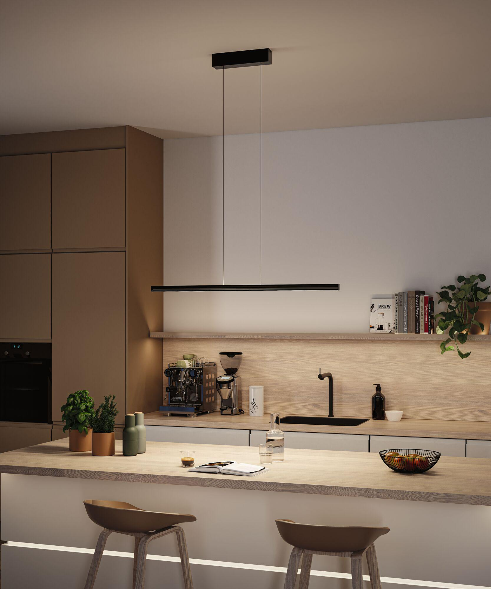 Nowoczesna kuchnia z barem i stołkami. Nad nim wisi czarna lampa. Ekspres do kawy, rośliny i miska owoców na blacie.