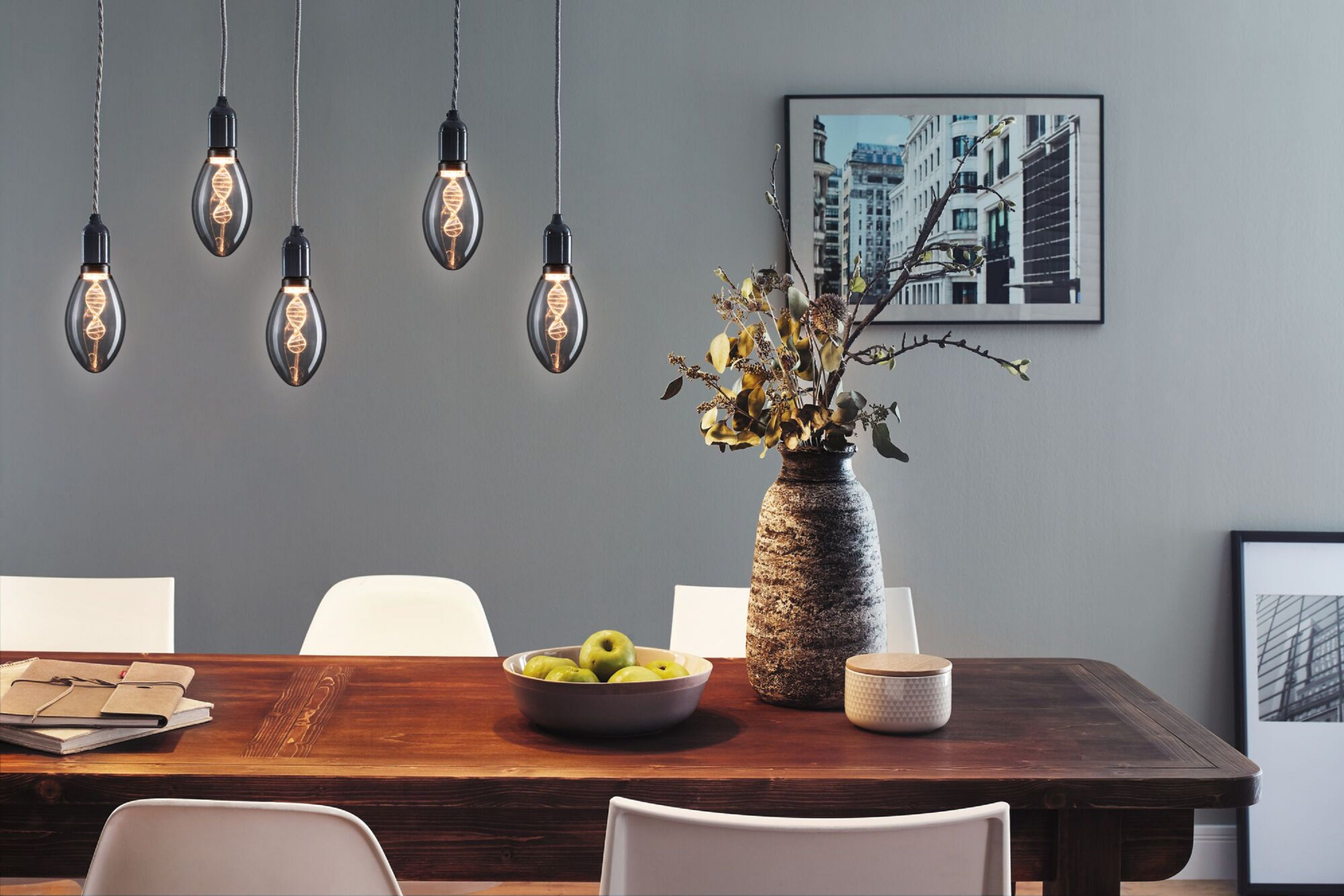 Esszimmer mit Hängelampen, einem Holztisch mit Äpfeln, einer Vase mit Blumen und gerahmten Bildern an der Wand.