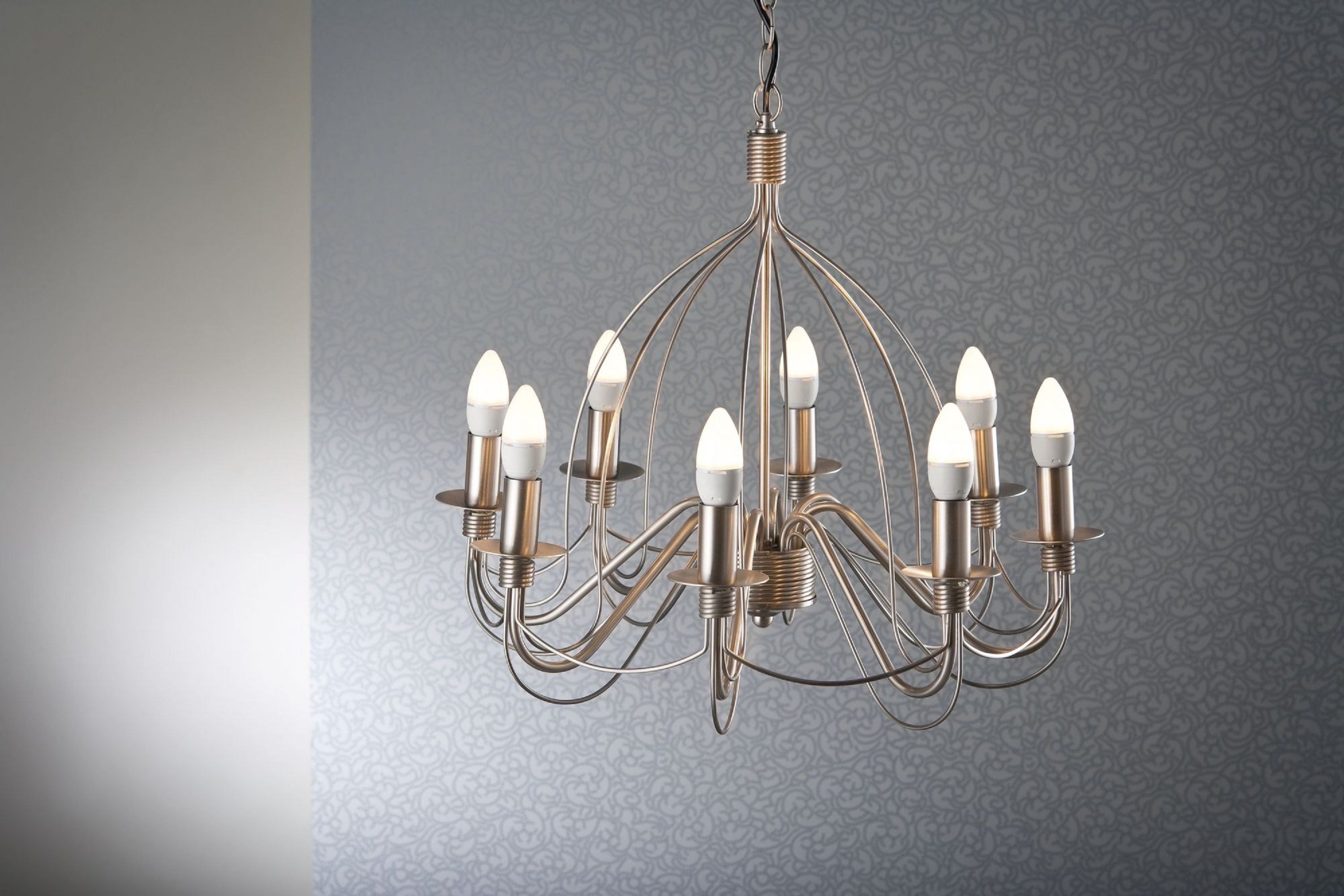 Un lampadario con dieci lampadine pende su uno sfondo grigio a motivi. La luce è accesa. Il lampadario è dorato.