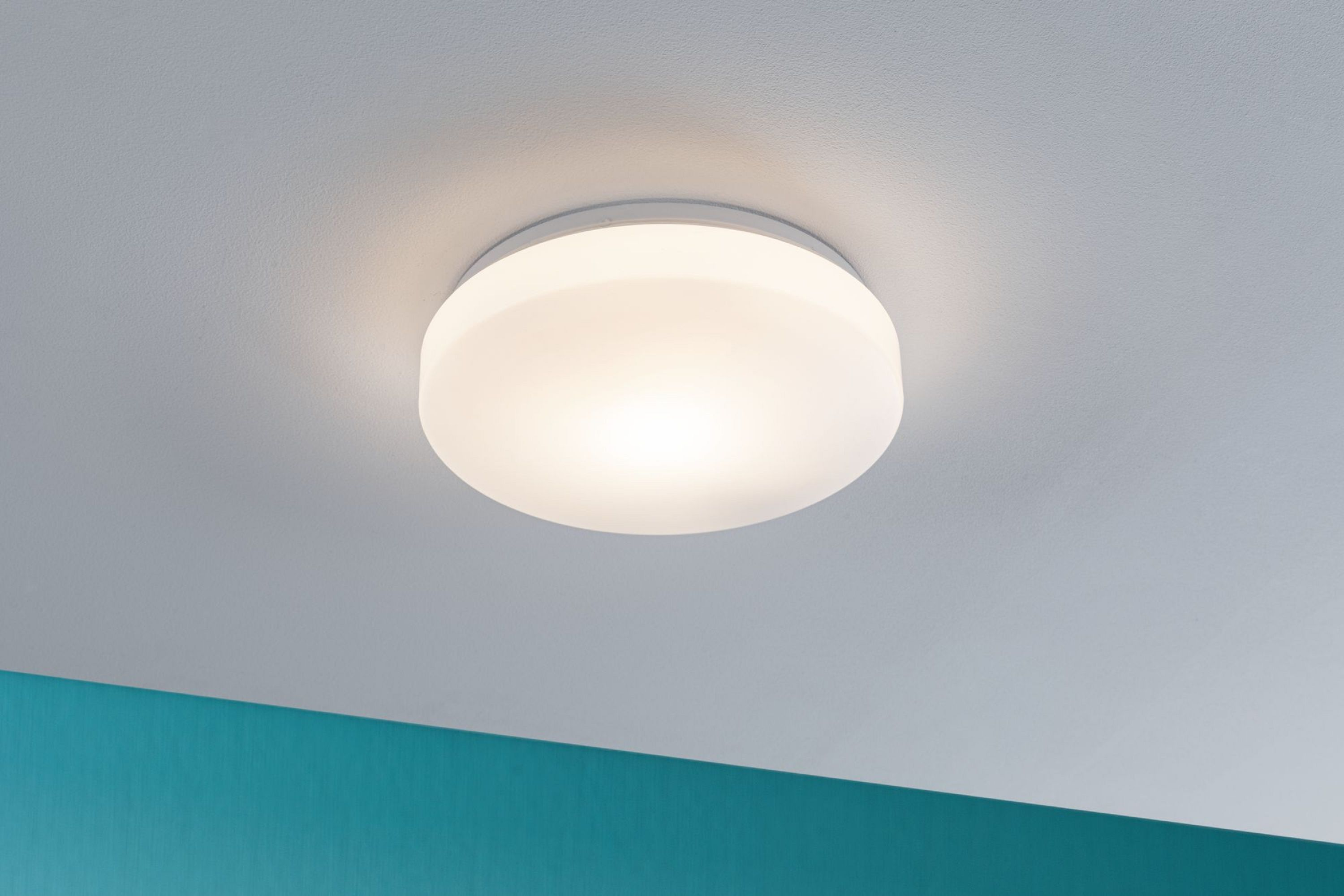 Un plafonnier rond et blanc est centré sur un plafond blanc. Un mur turquoise est en bas à gauche.